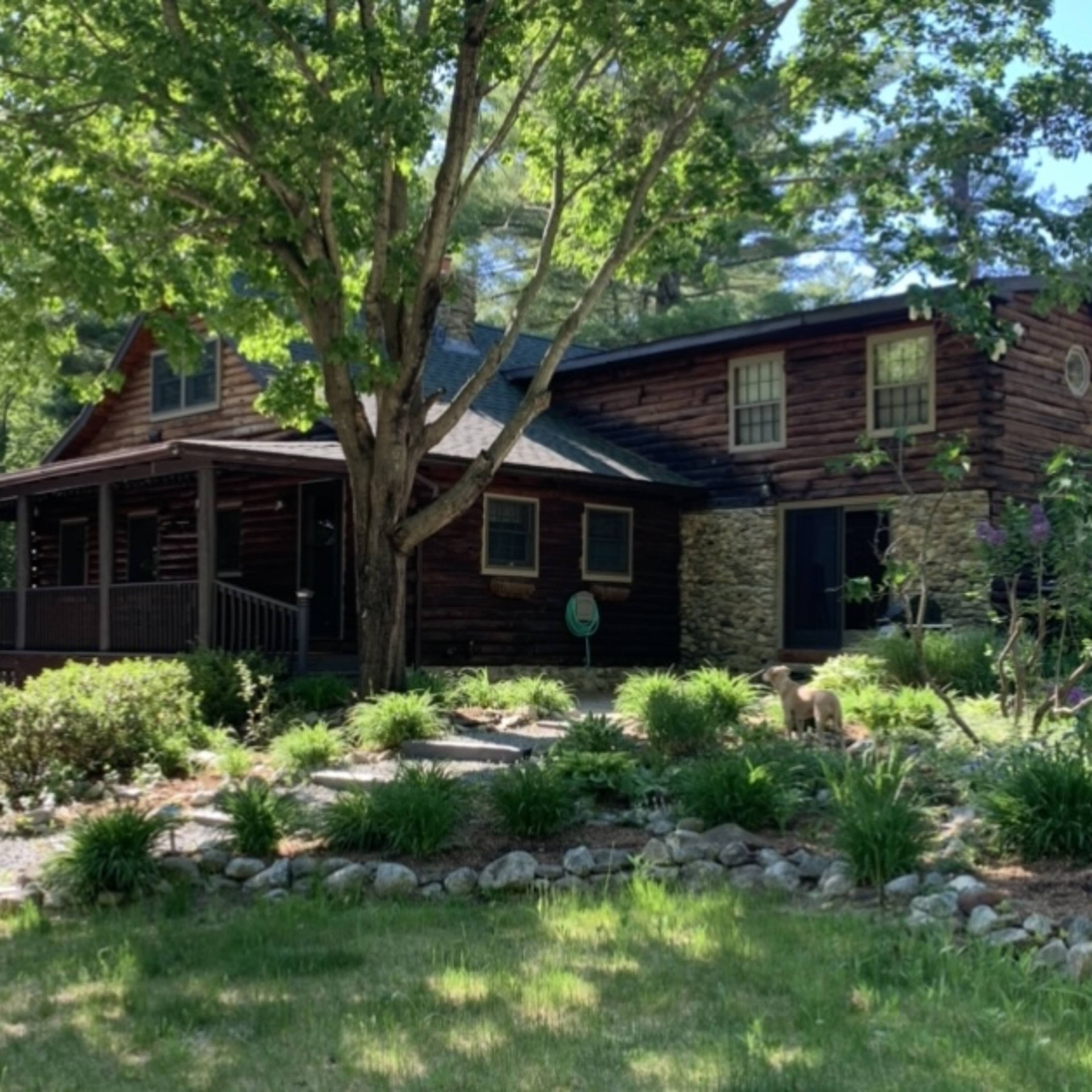 A wooden house with a stone section and a porch is surrounded by greenery and trees.