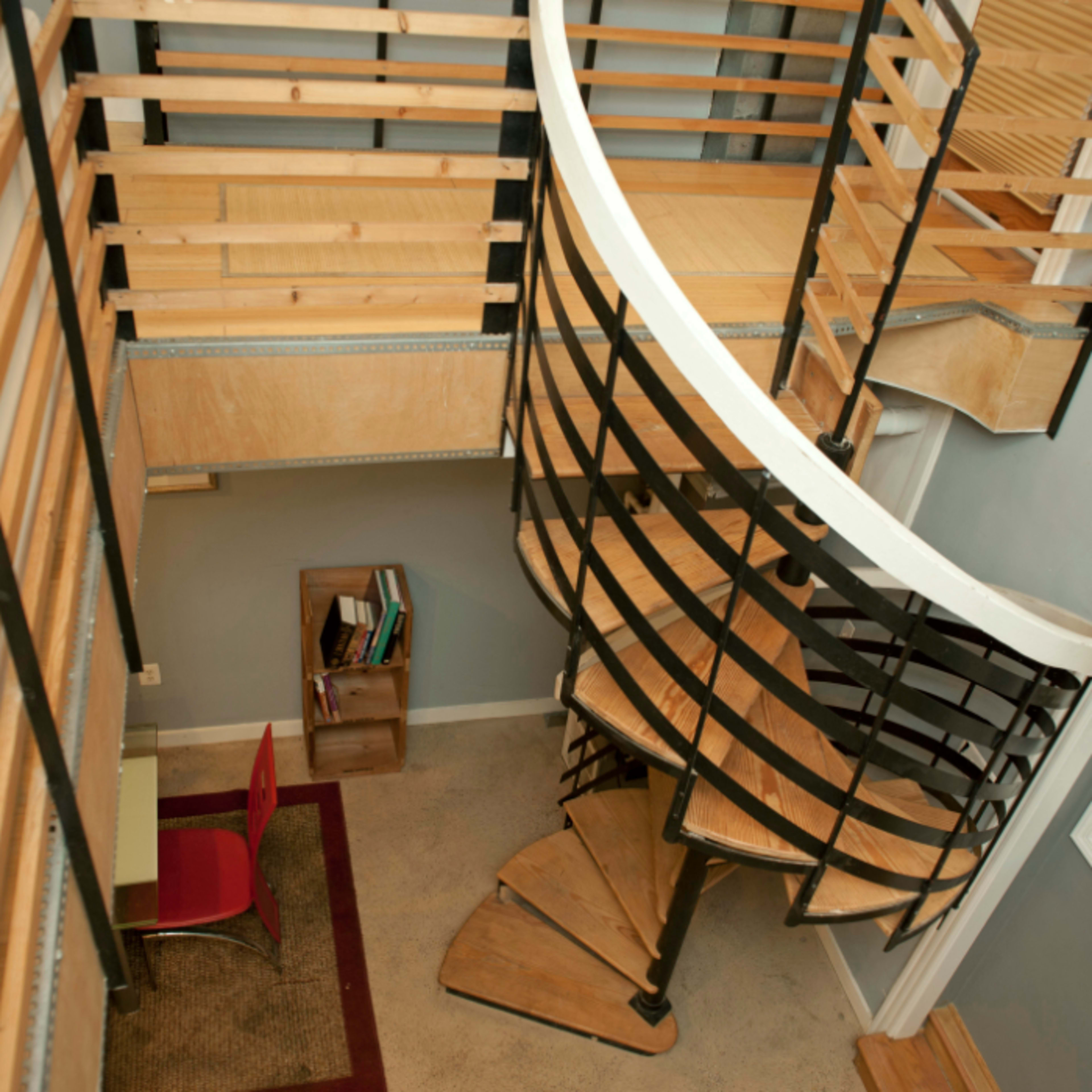 A two-story interior space featuring a curved staircase, wooden railings, and a desk area with a red chair.