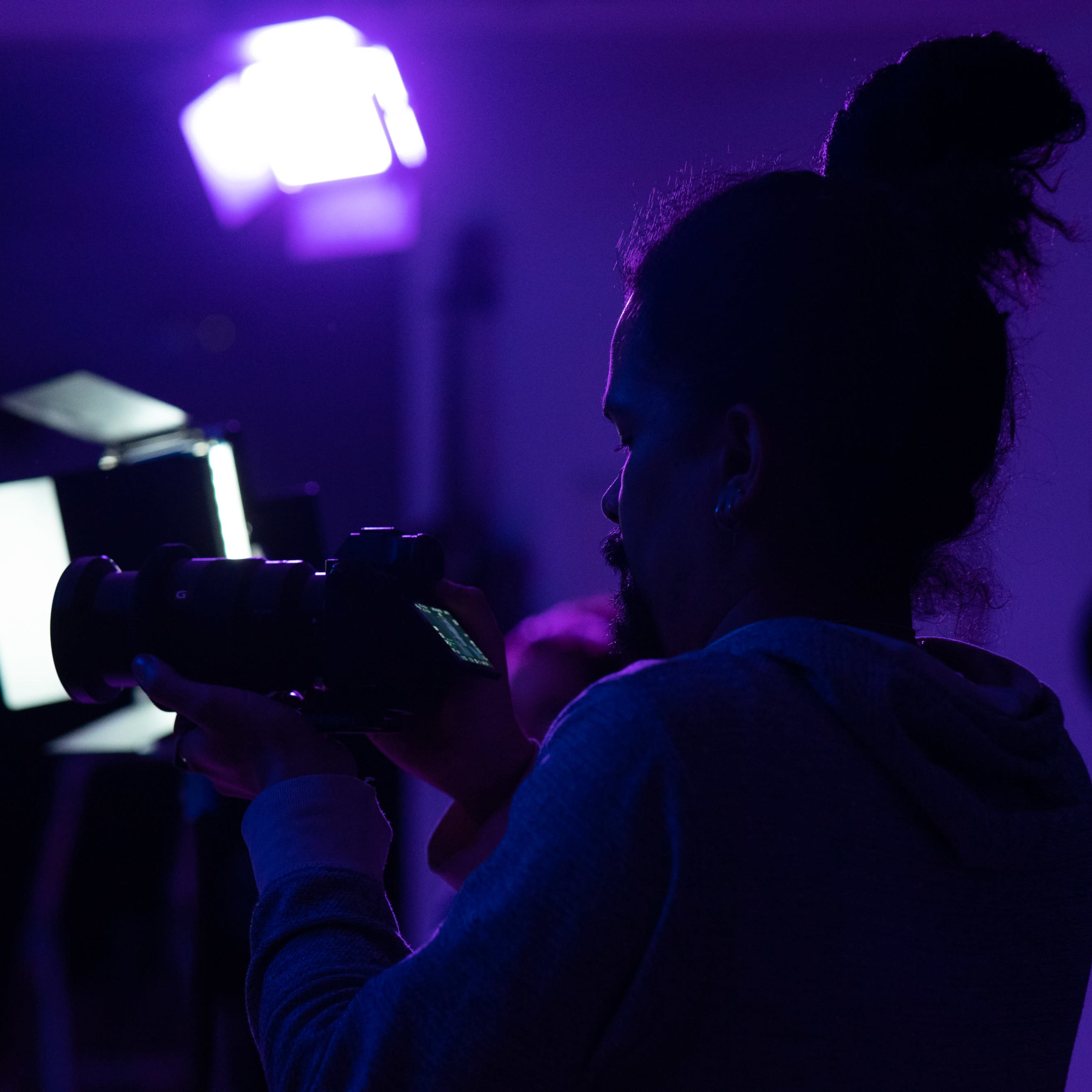 A person holds a camera while standing in a dimly lit room with purple lighting and guitars visible in the background.