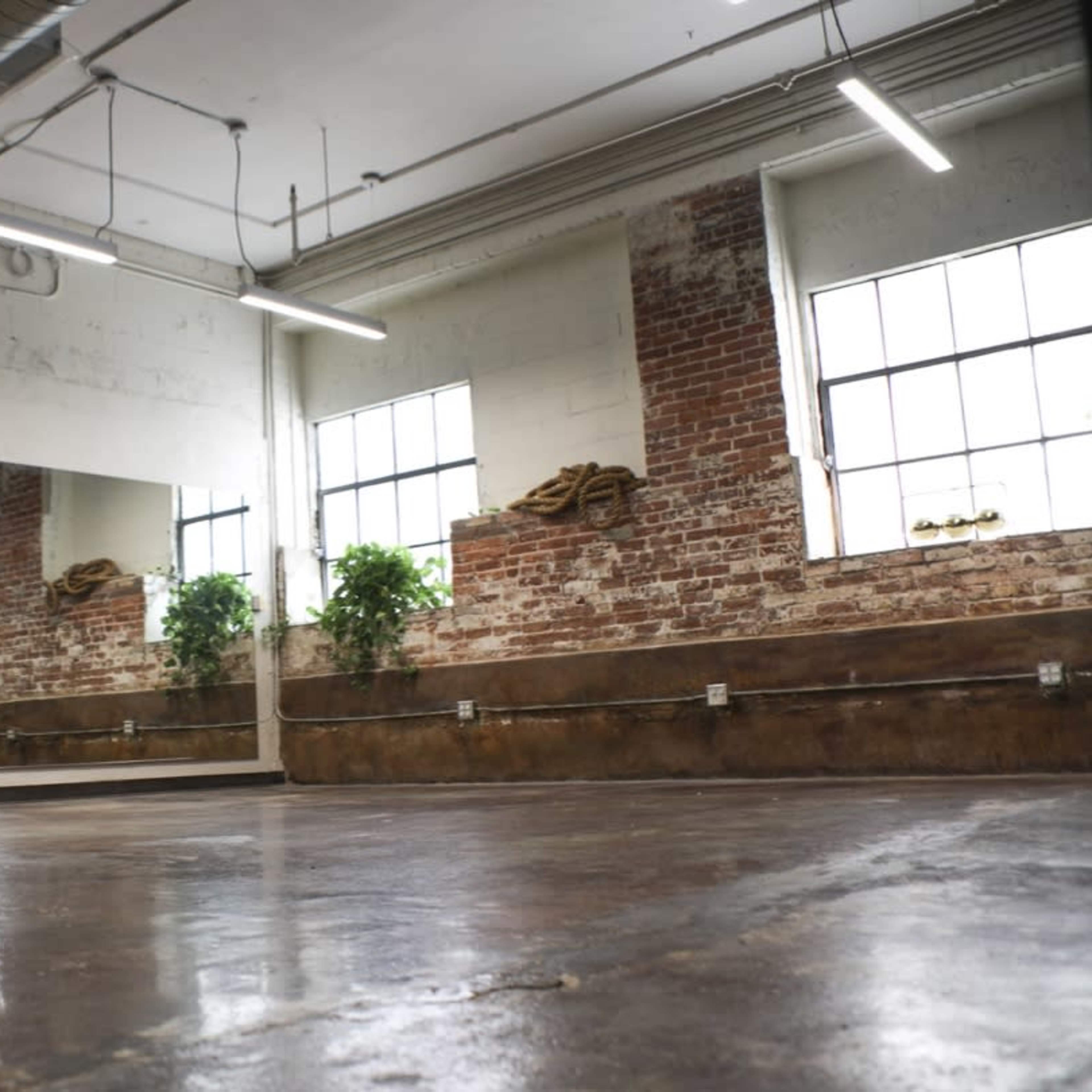 An empty industrial-style room with exposed brick walls, large windows, and a polished concrete floor, featuring a mirror and potted plants.