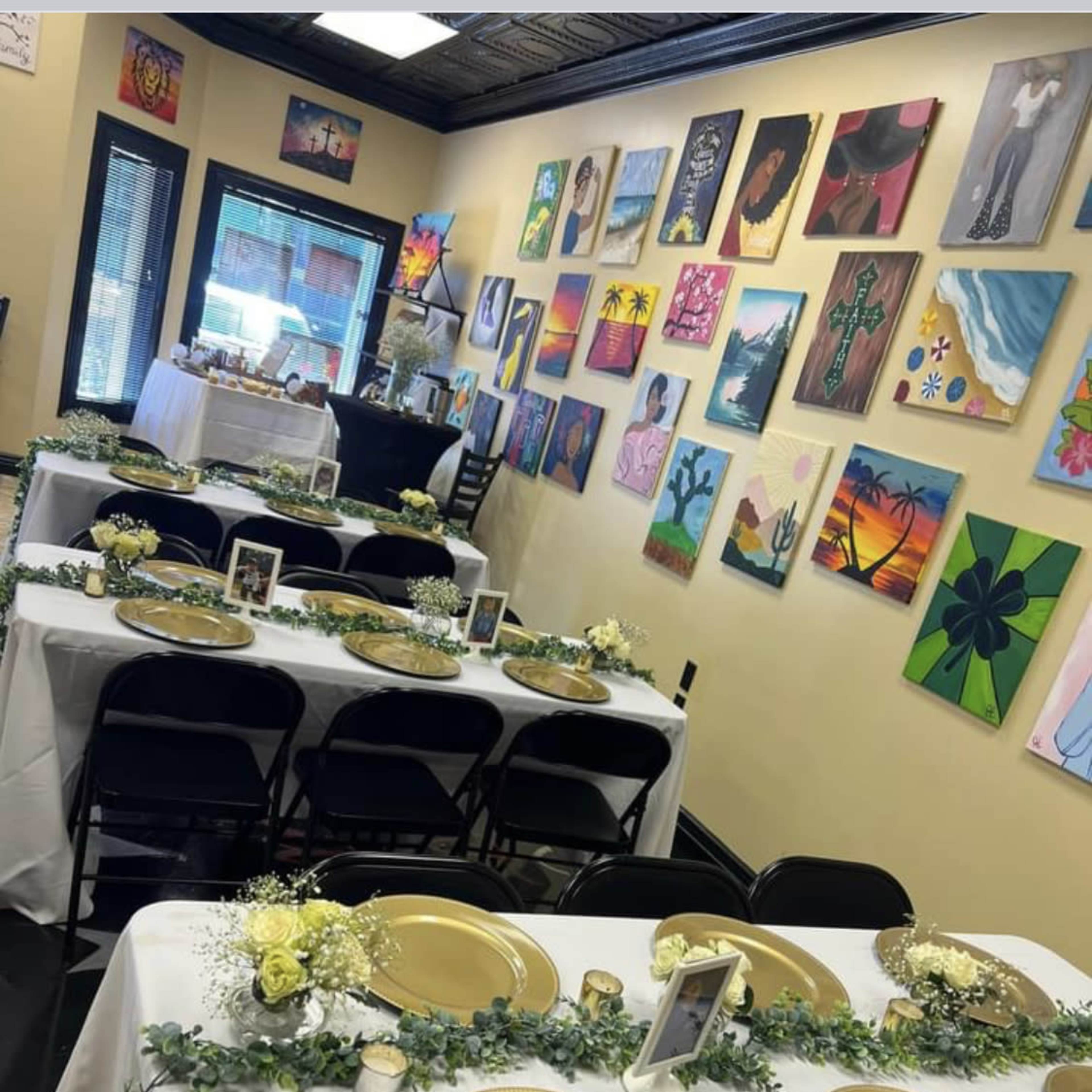 A decorated dining area with multiple tables set for an event, featuring gold plates and floral centerpieces against a backdrop of colorful artwork on the walls.