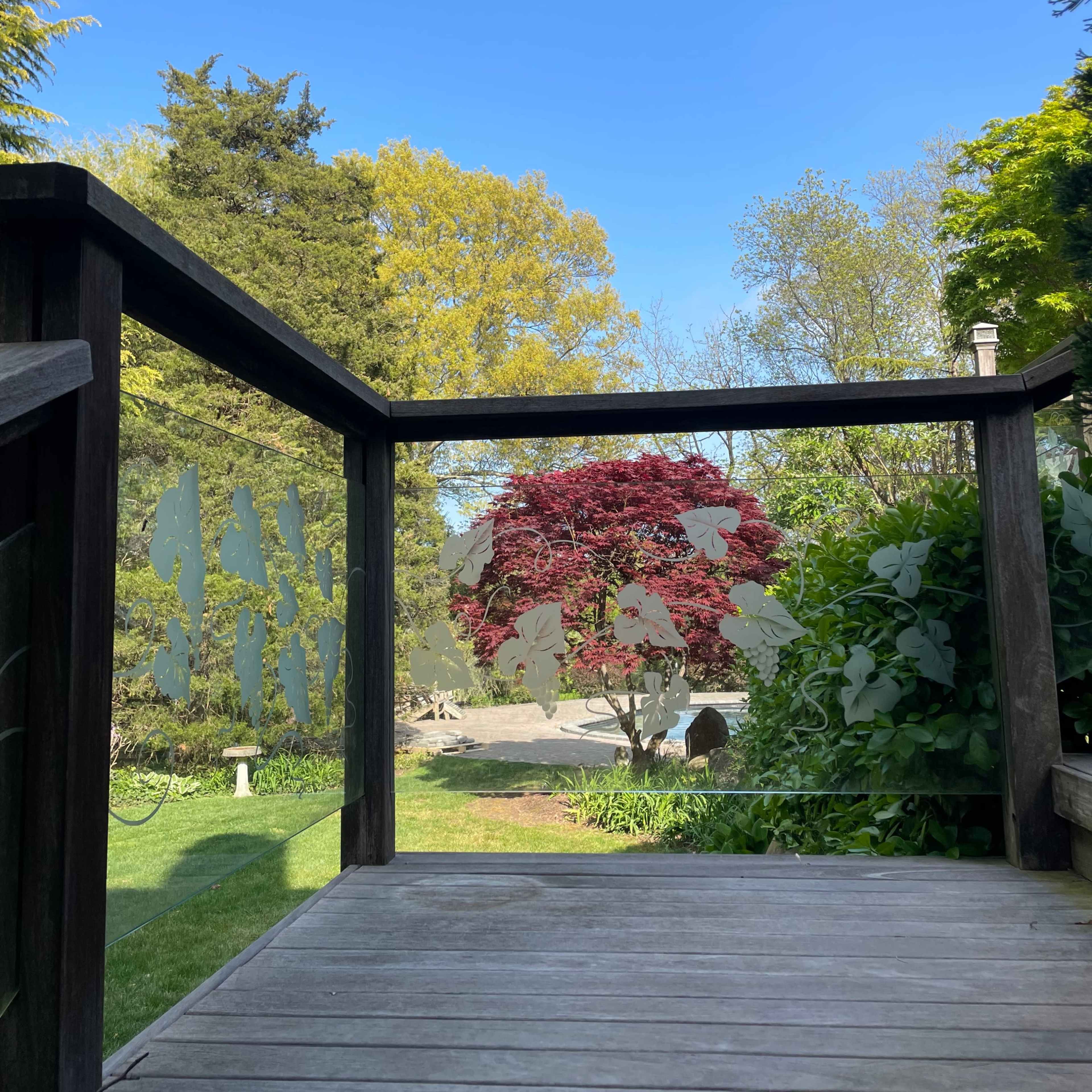 A wooden deck with a glass railing overlooks a well-maintained garden featuring a vibrant red tree and greenery under a clear blue sky.