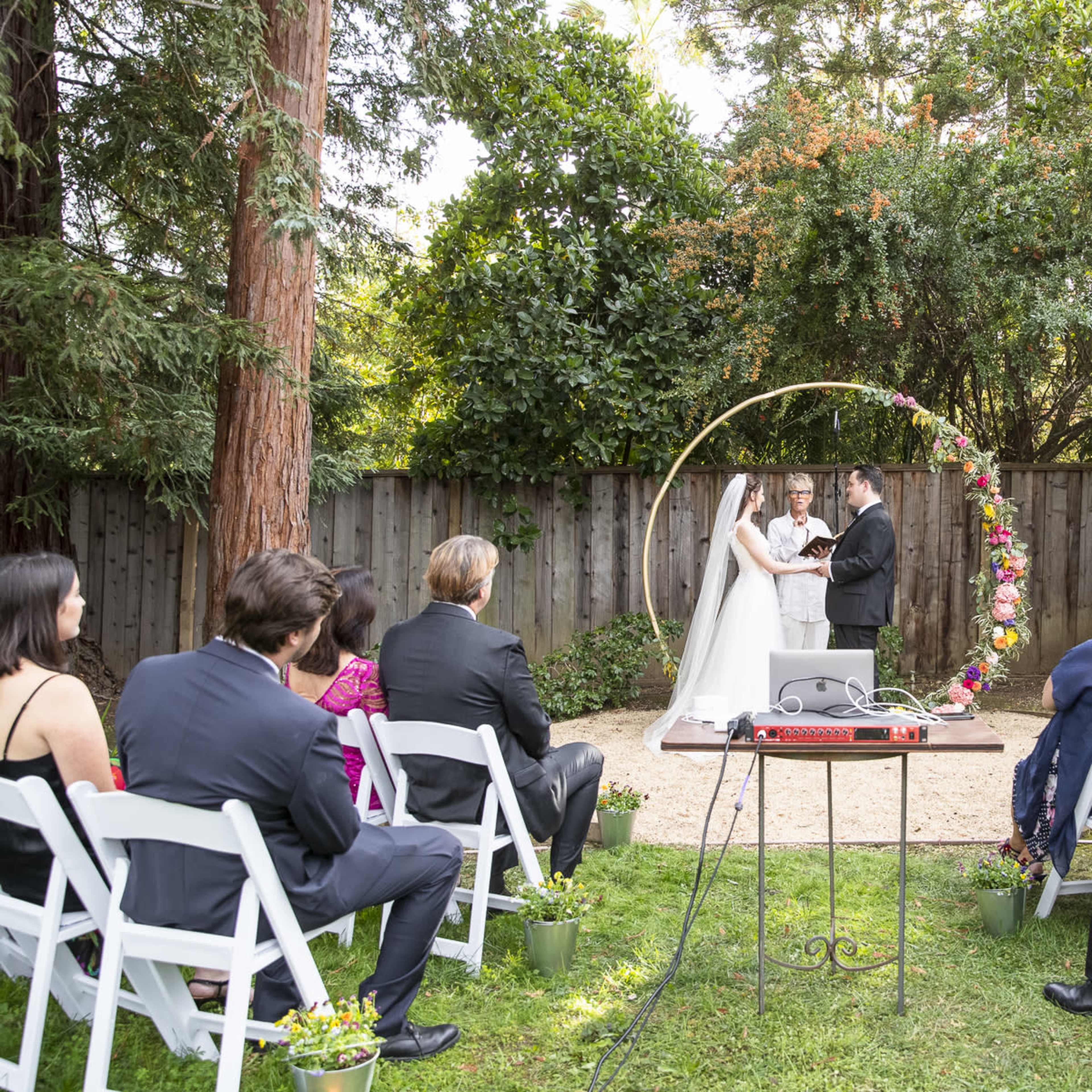 A couple exchanges vows under a floral arch while guests sit on white chairs in a garden setting.