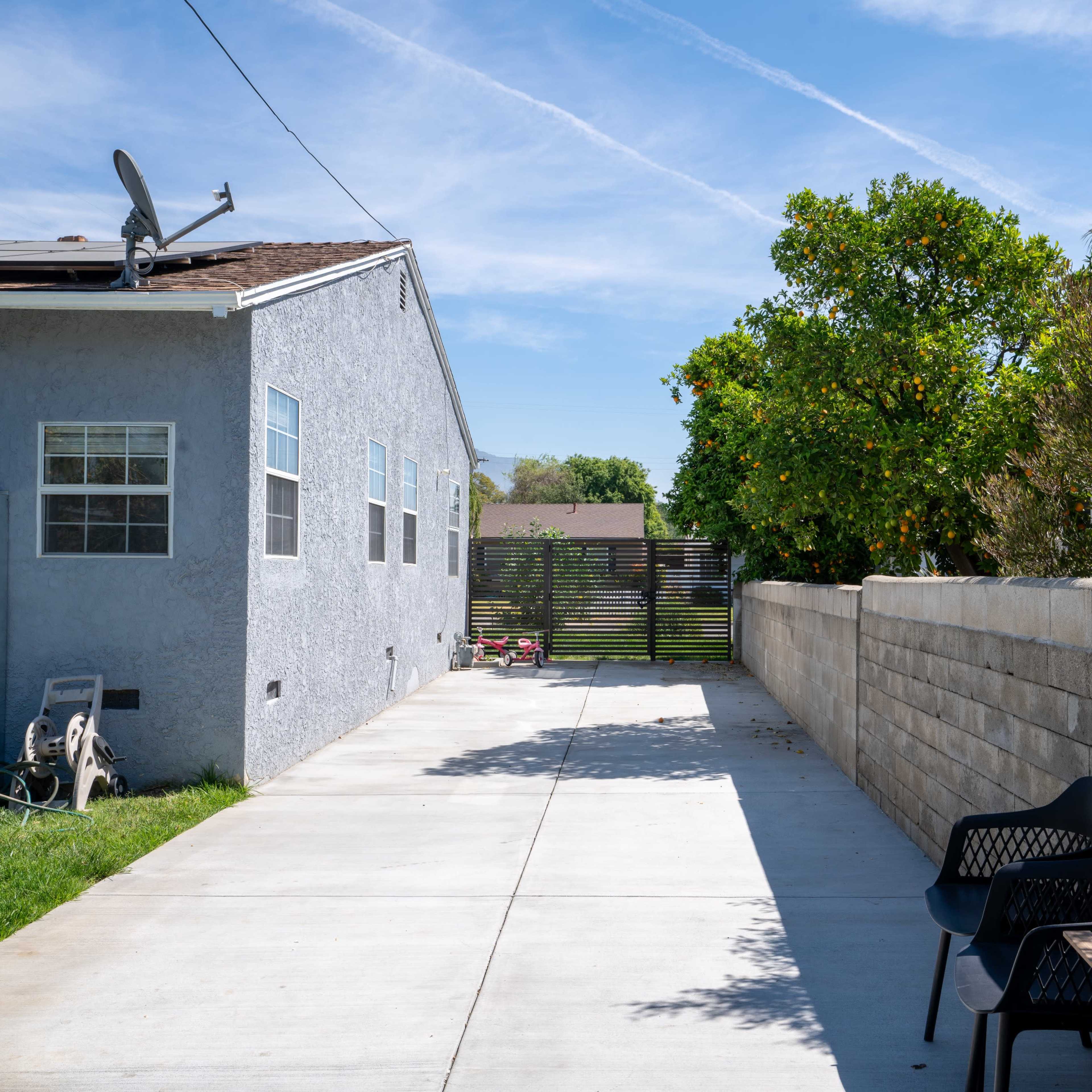 A concrete pathway leads alongside a gray house towards a gated entrance, bordered by a stone wall and a grassy area.