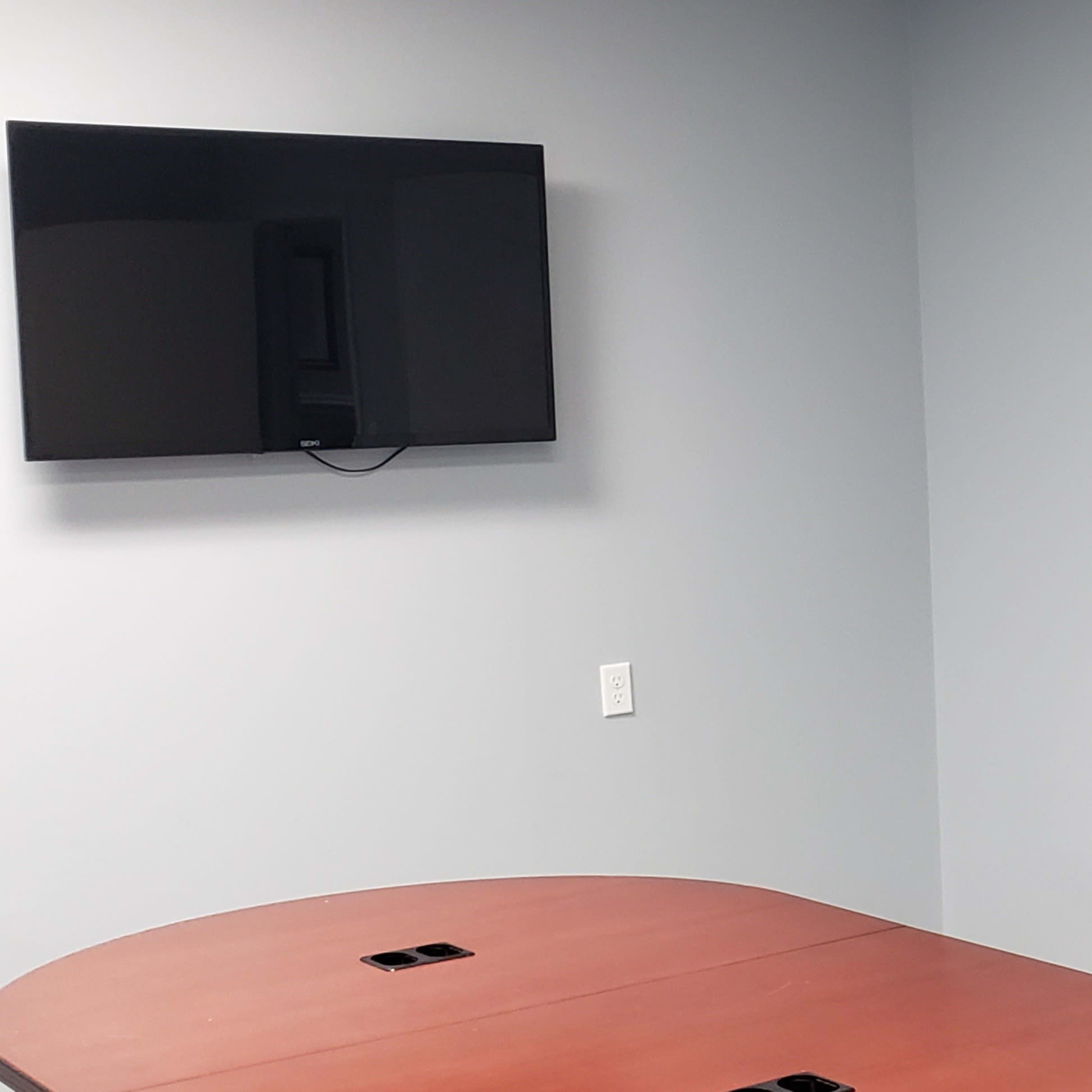 A meeting room with a brown table, a wall-mounted television, and a blank whiteboard against a gray wall.