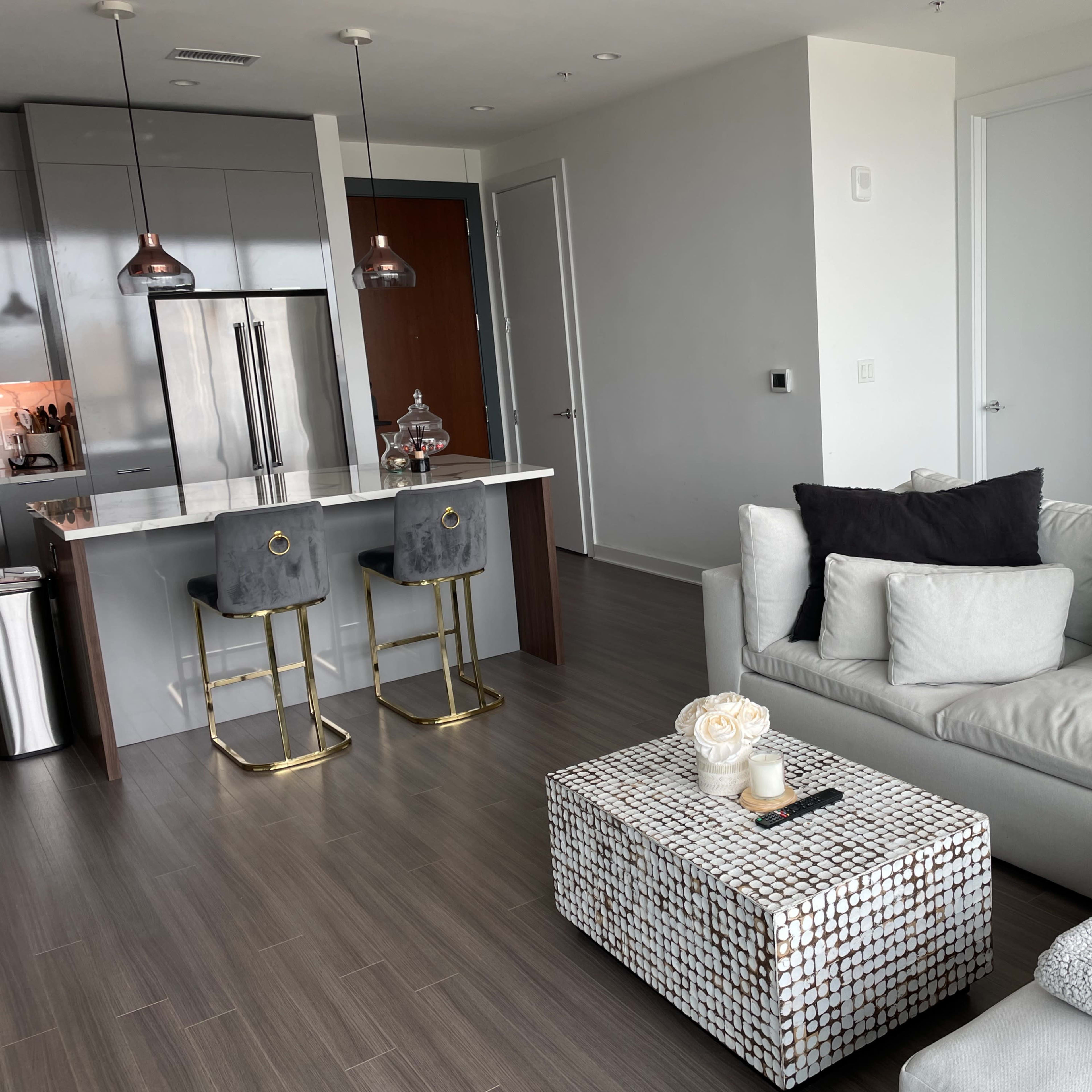 A modern living room features a gray sectional couch, a mirrored coffee table, and a kitchen area with bar stools and stainless steel appliances.