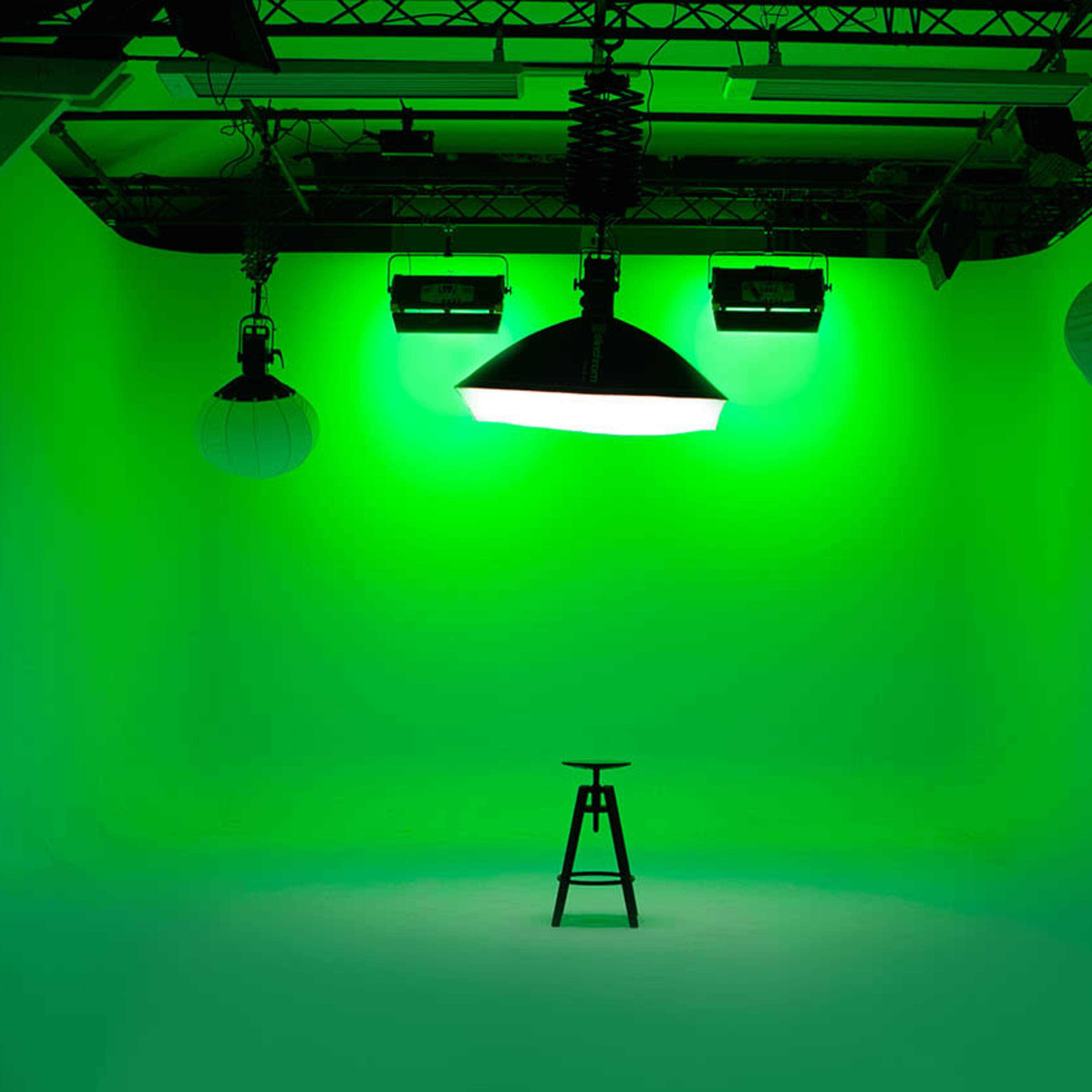 A photography studio features a green backdrop with various lighting fixtures and a single stool in the center.