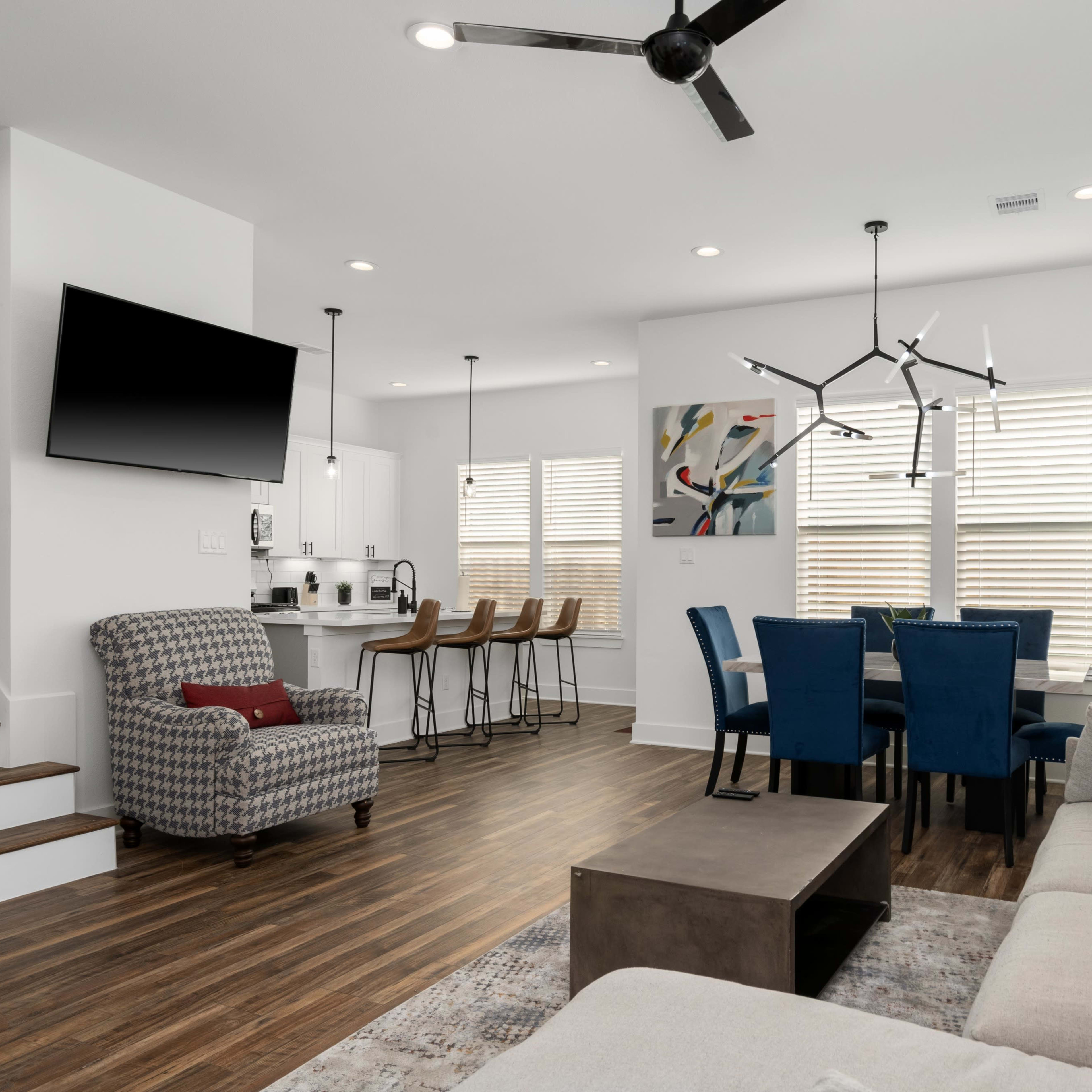 A modern living room and dining area featuring a sofa, a coffee table, a television mounted on the wall, and a dining table with blue chairs.