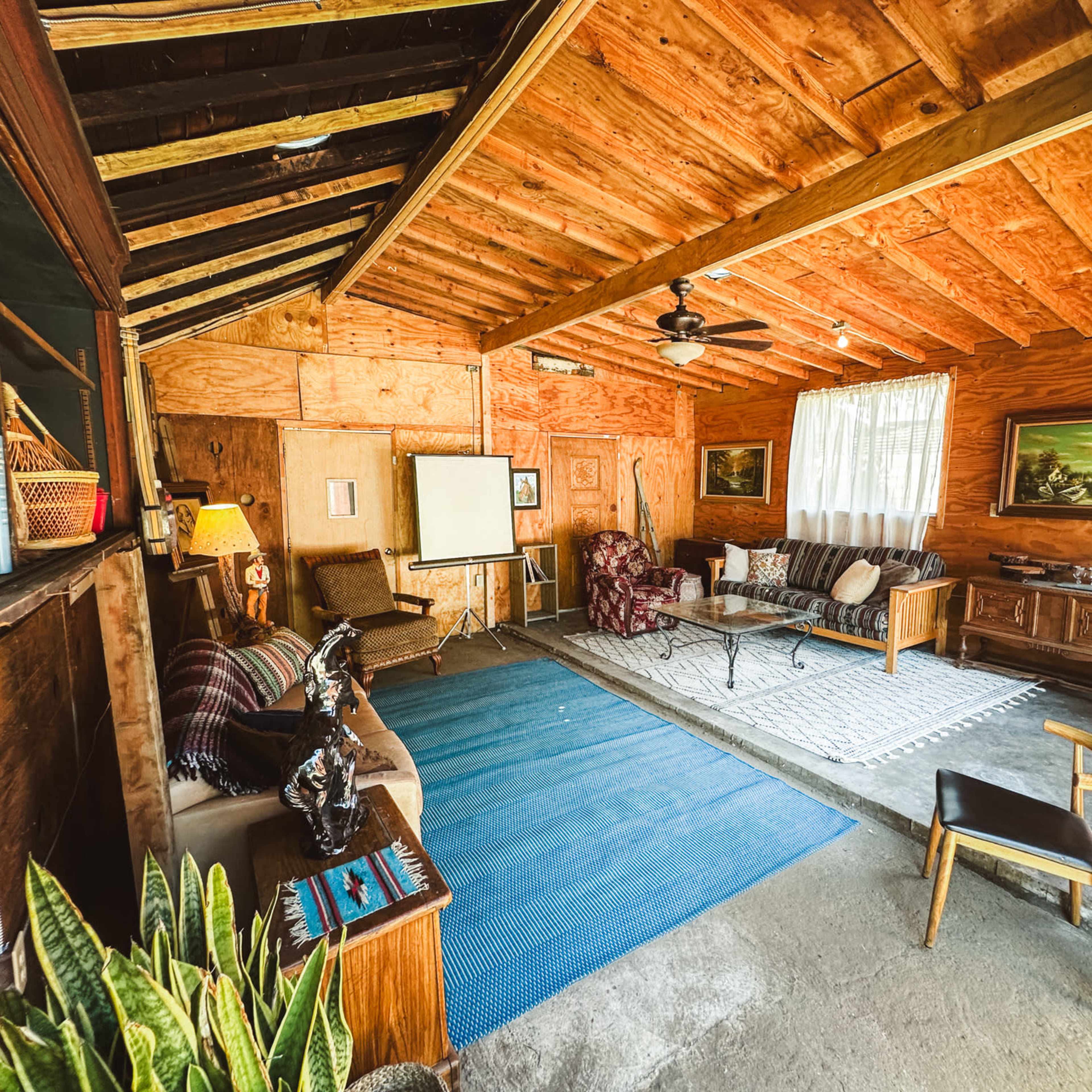 The interior of a rustic cabin features wooden walls, a ceiling fan, a seating area with various furniture, and a large window letting in natural light.