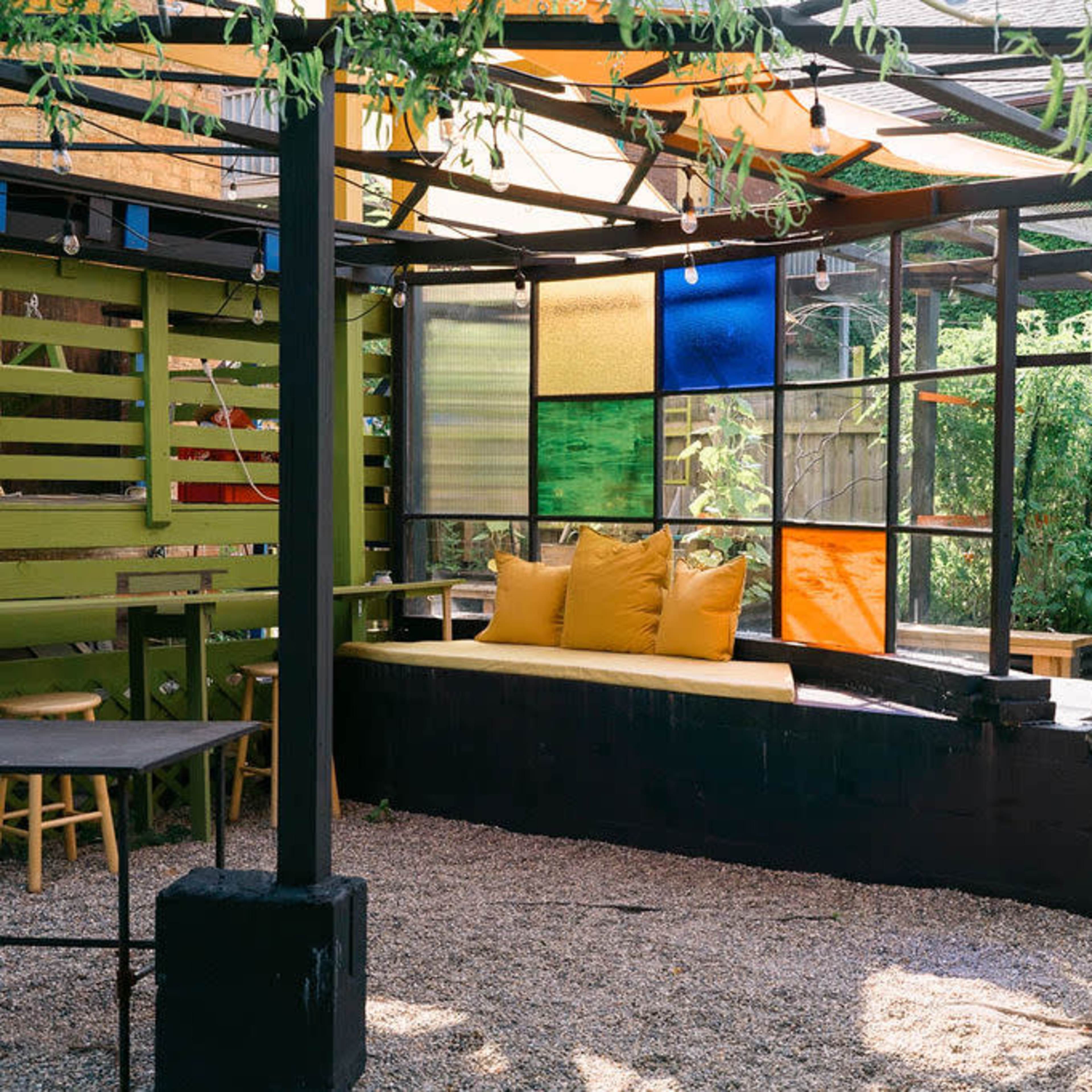 A modern outdoor seating area features a large, colorful glass panel wall, a yellow cushioned bench, and gravel flooring surrounded by greenery.