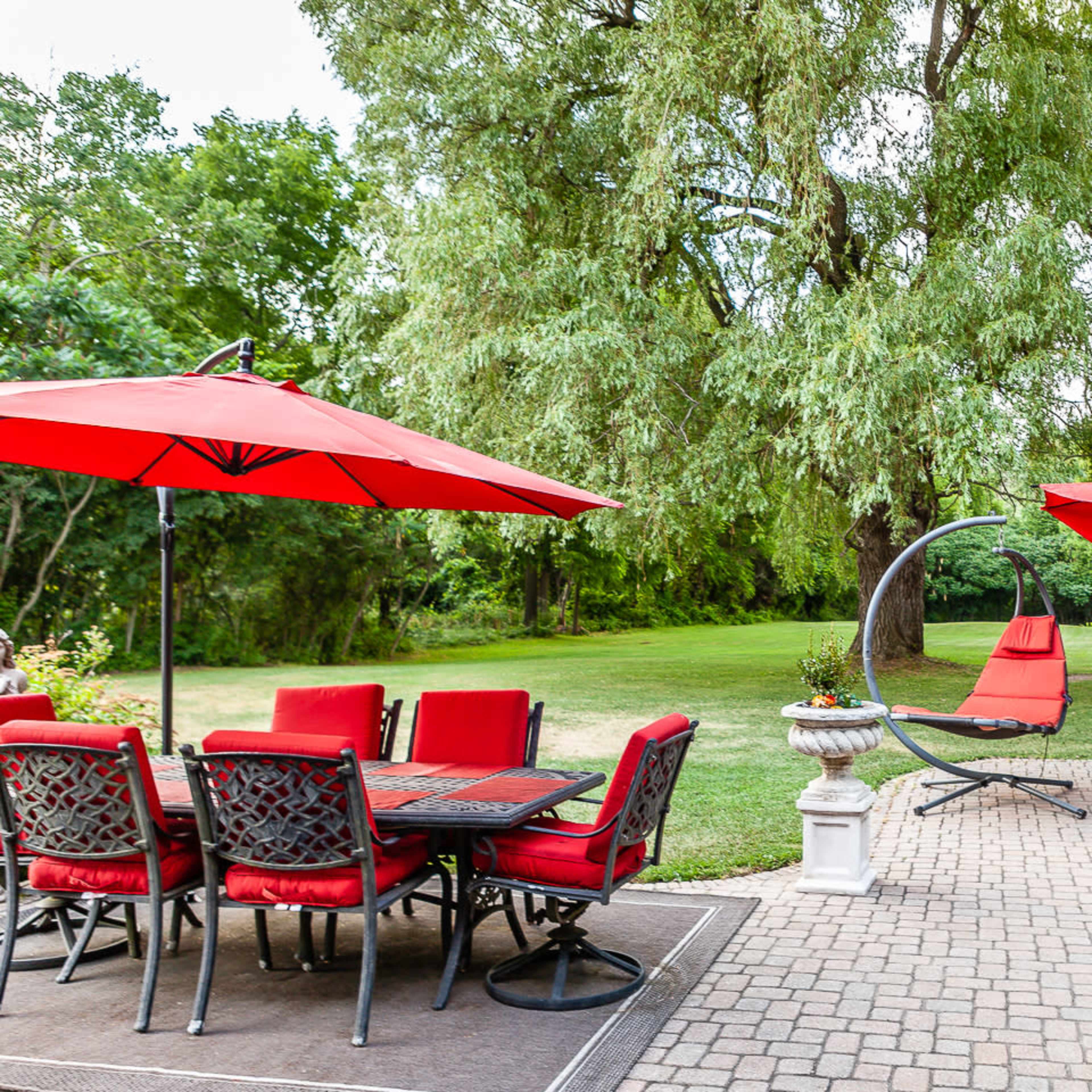 A backyard patio with a hot tub, seating area, and a dining set under red umbrellas, surrounded by greenery.