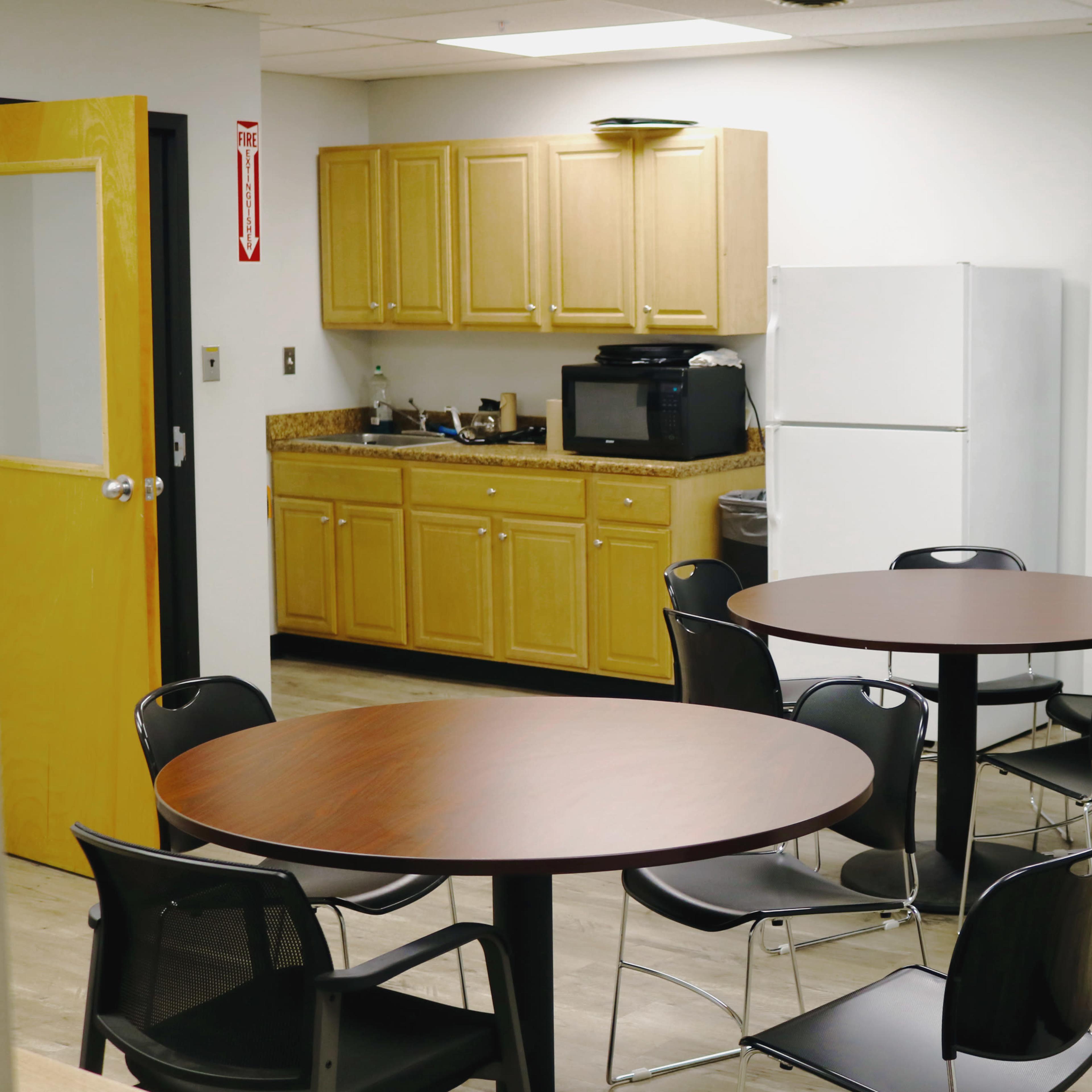 A break room with two circular tables and black chairs, a kitchenette with wooden cabinets, a microwave, and a refrigerator against the wall.