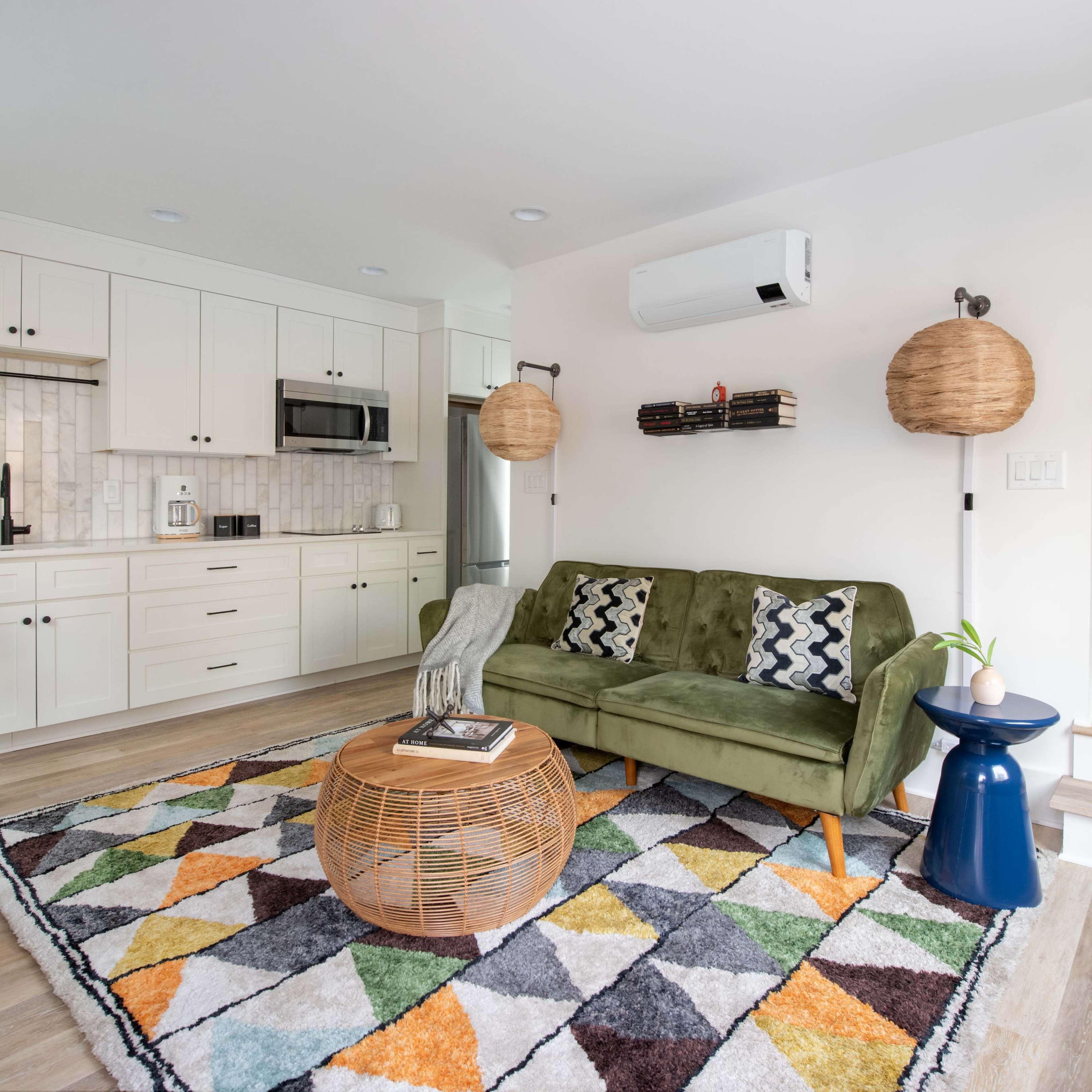 A modern living space featuring a green sofa, a patterned rug, a wooden coffee table, and a kitchen area with white cabinets.