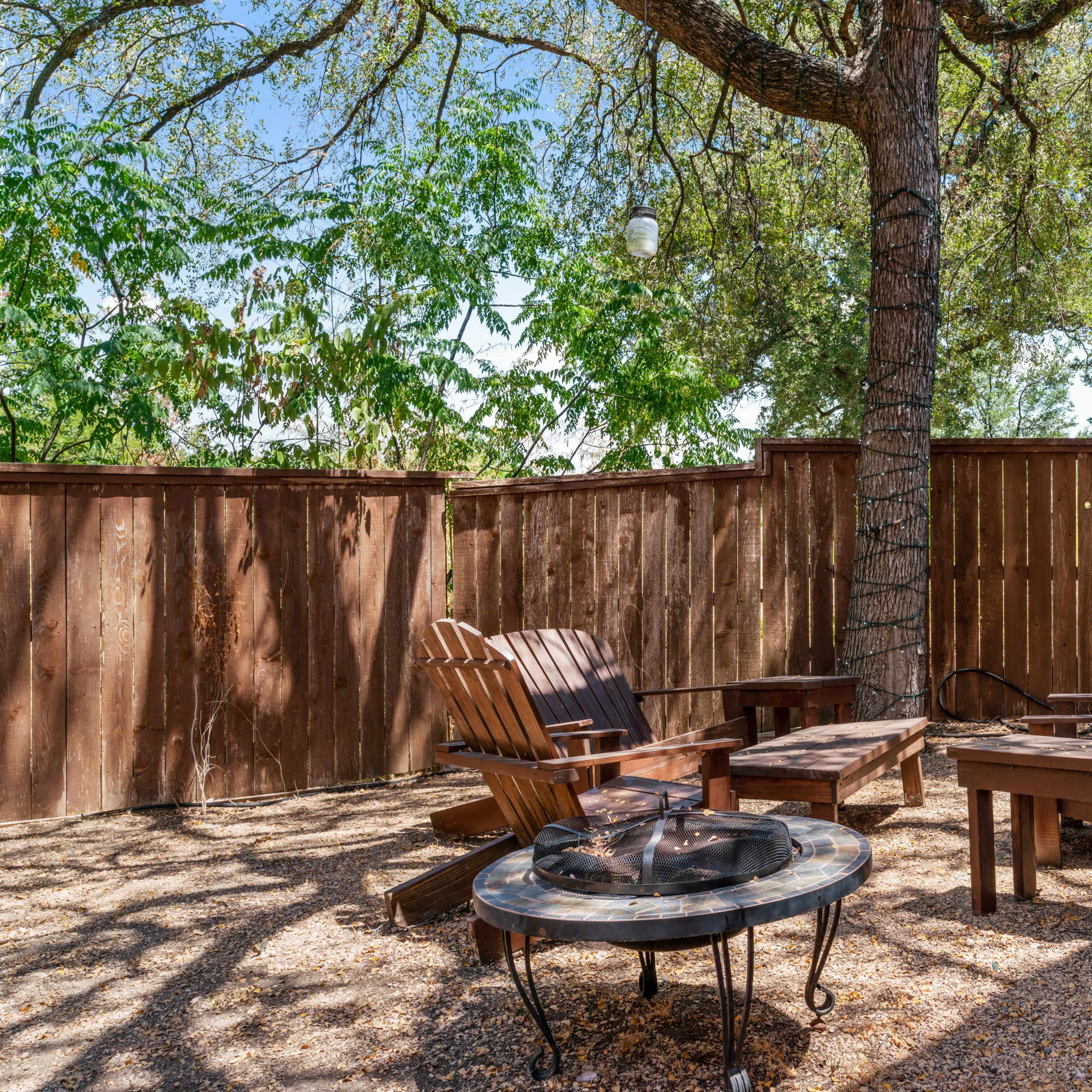 A seating area with wooden chairs and a fire pit is situated under a tree surrounded by a wooden fence.