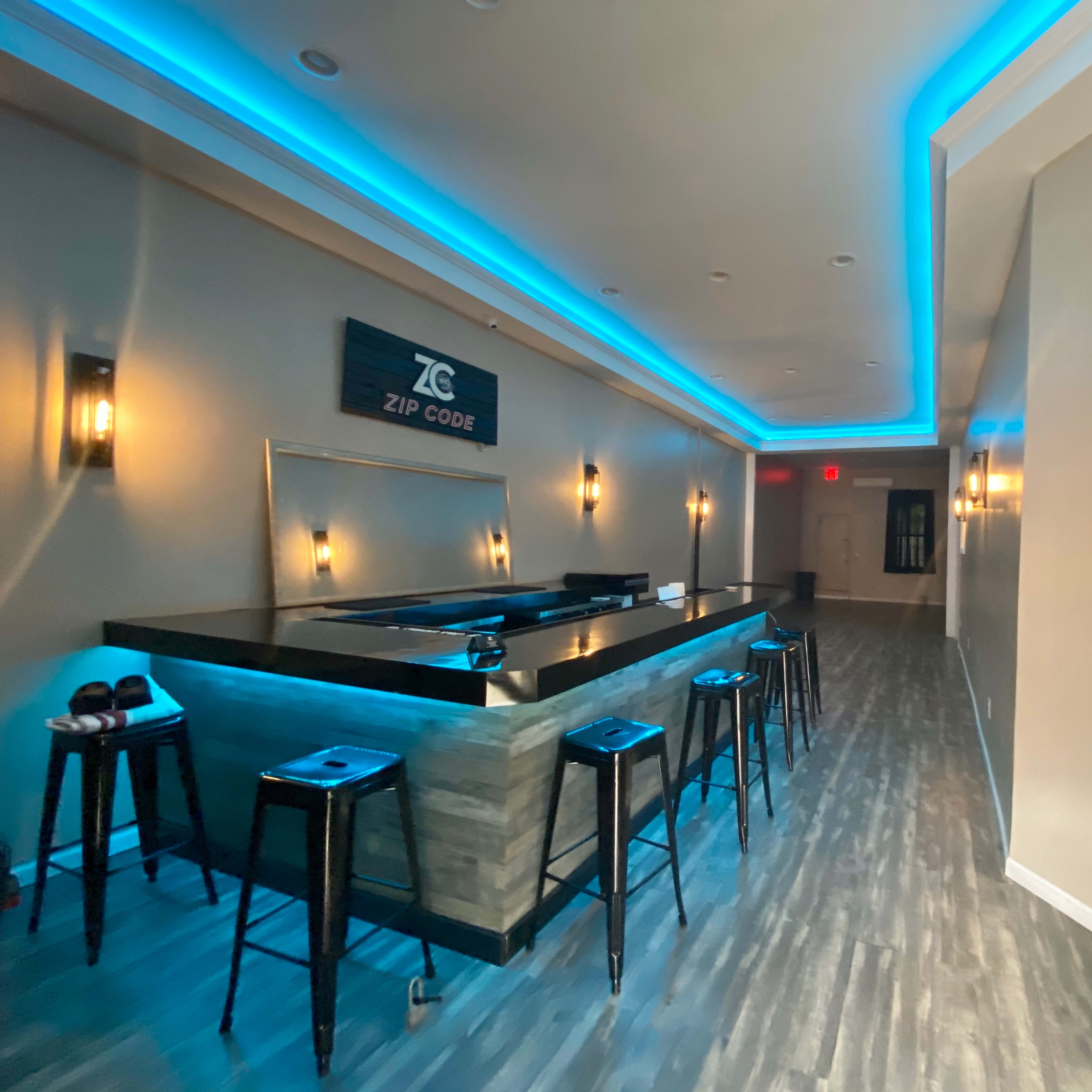 A modern bar area with a wooden counter and high stools, illuminated by blue LED lights along the ceiling and behind the bar.