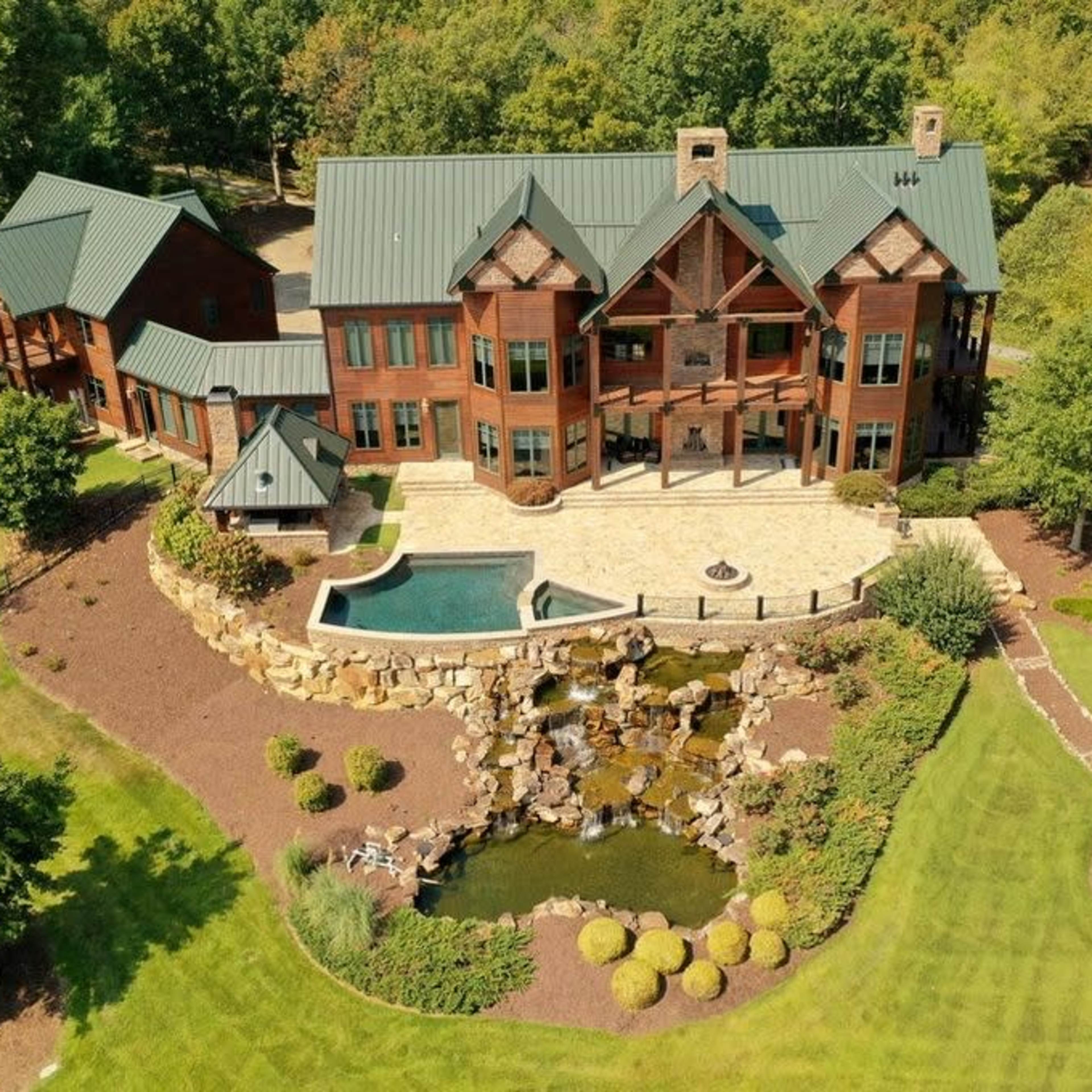 A large, multi-story wooden house with a green metal roof, surrounded by landscaped gardens and a pool with a cascading waterfall.