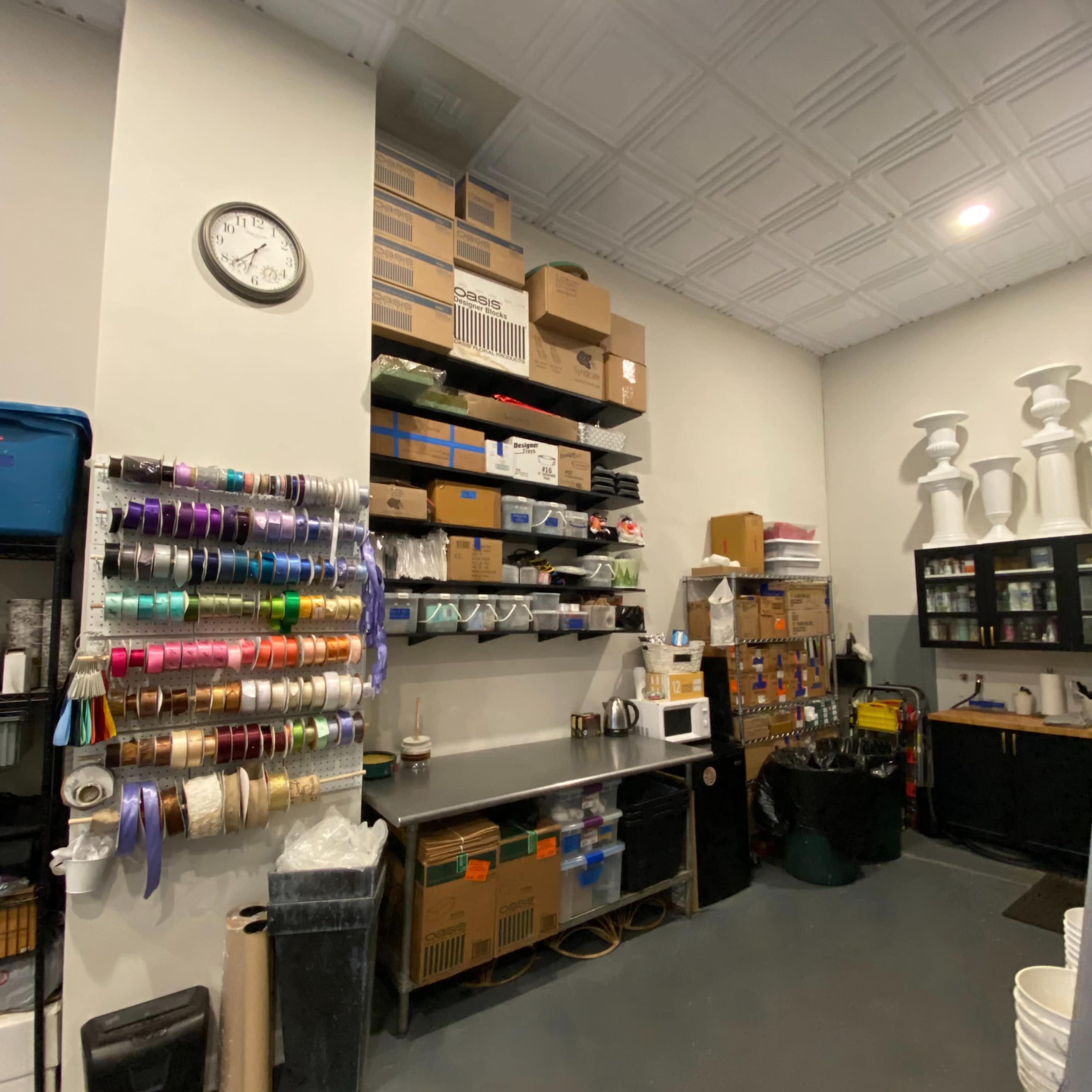 A storage area with organized shelves containing boxes, ribbons, and various supplies, alongside a clock on the wall and a small kitchenette.