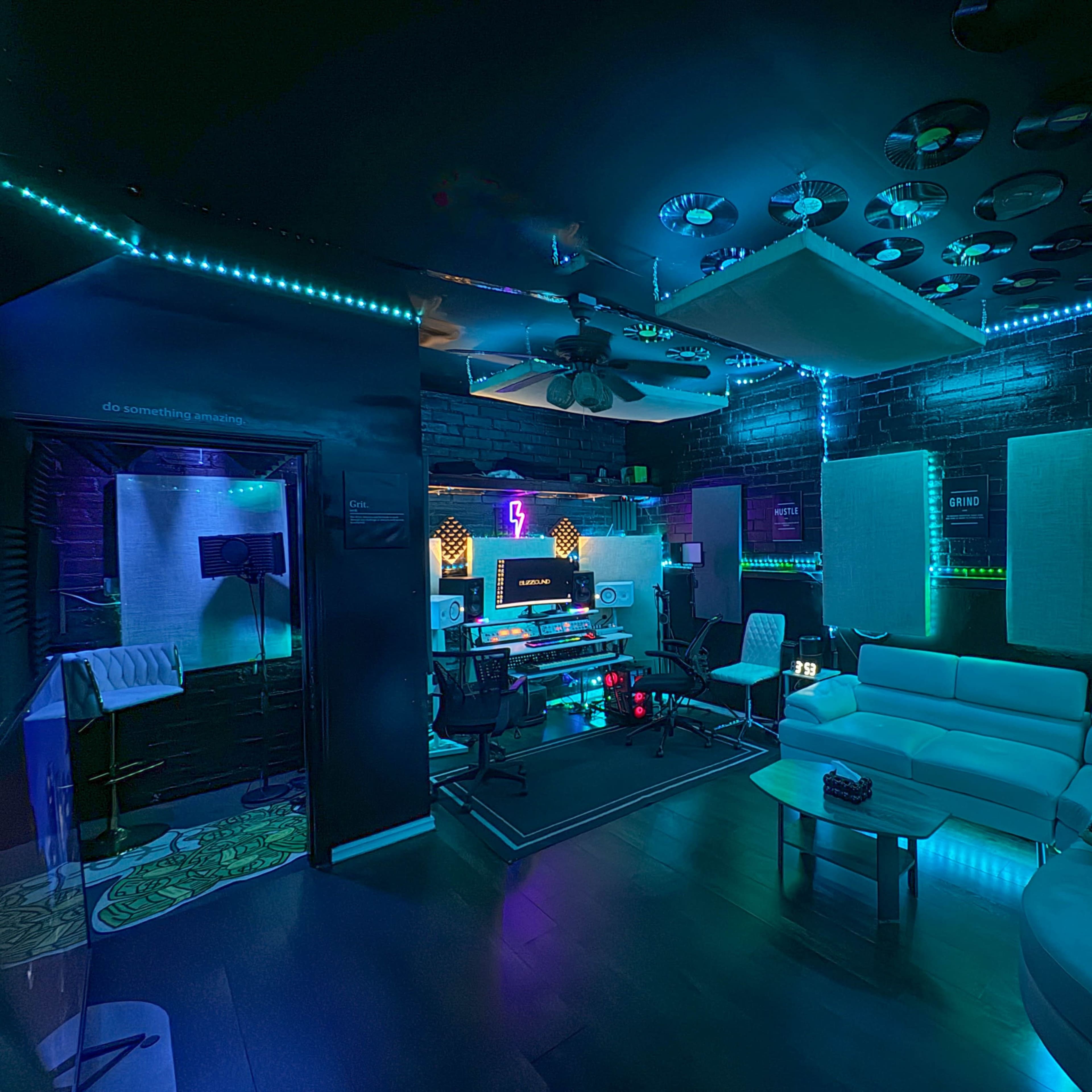 A music production studio features a mix of modern equipment, a white sofa, and ambient LED lighting against dark walls.