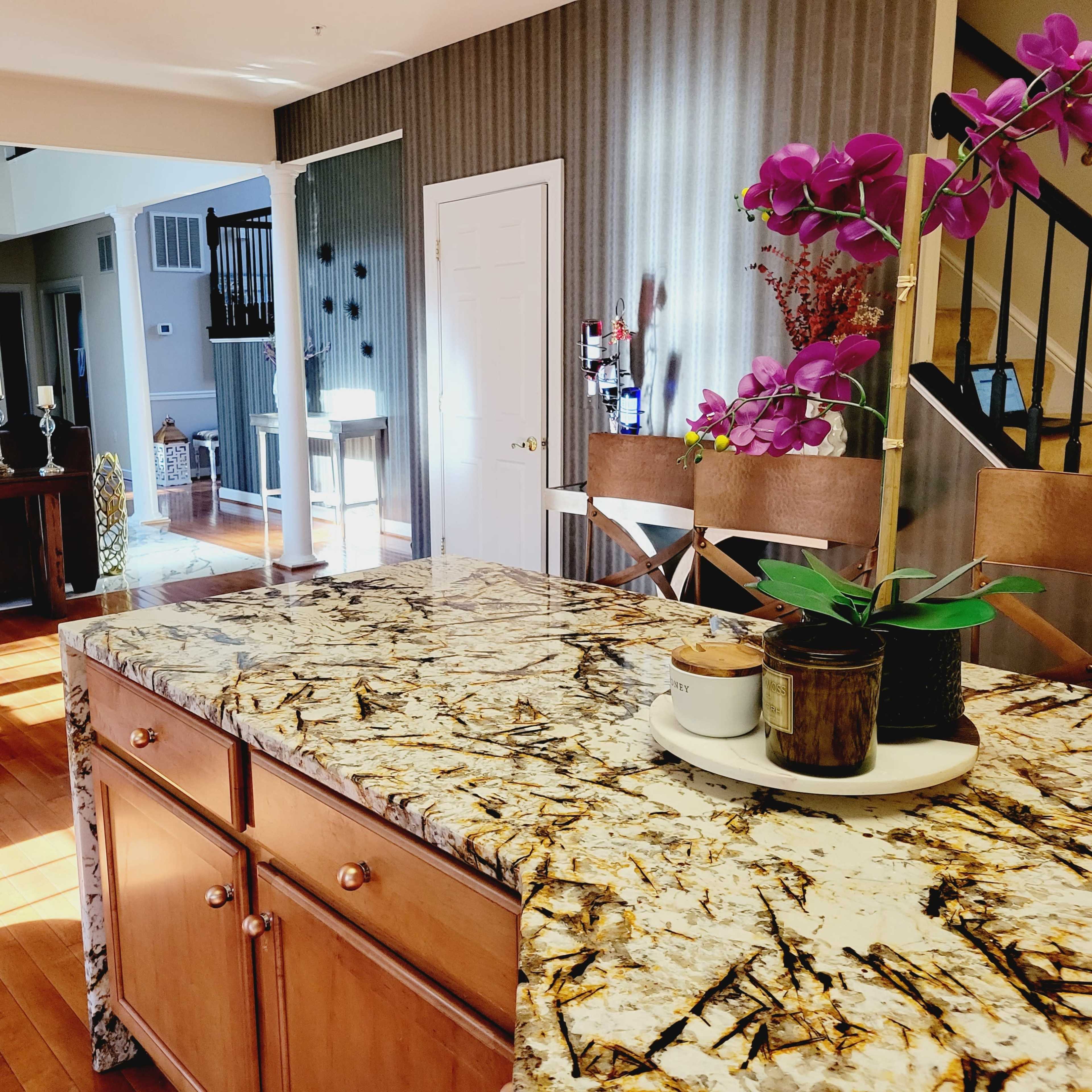 A bright kitchen with a marble countertop, wooden cabinets, and a view into a living area furnished with a sofa and decorative items.