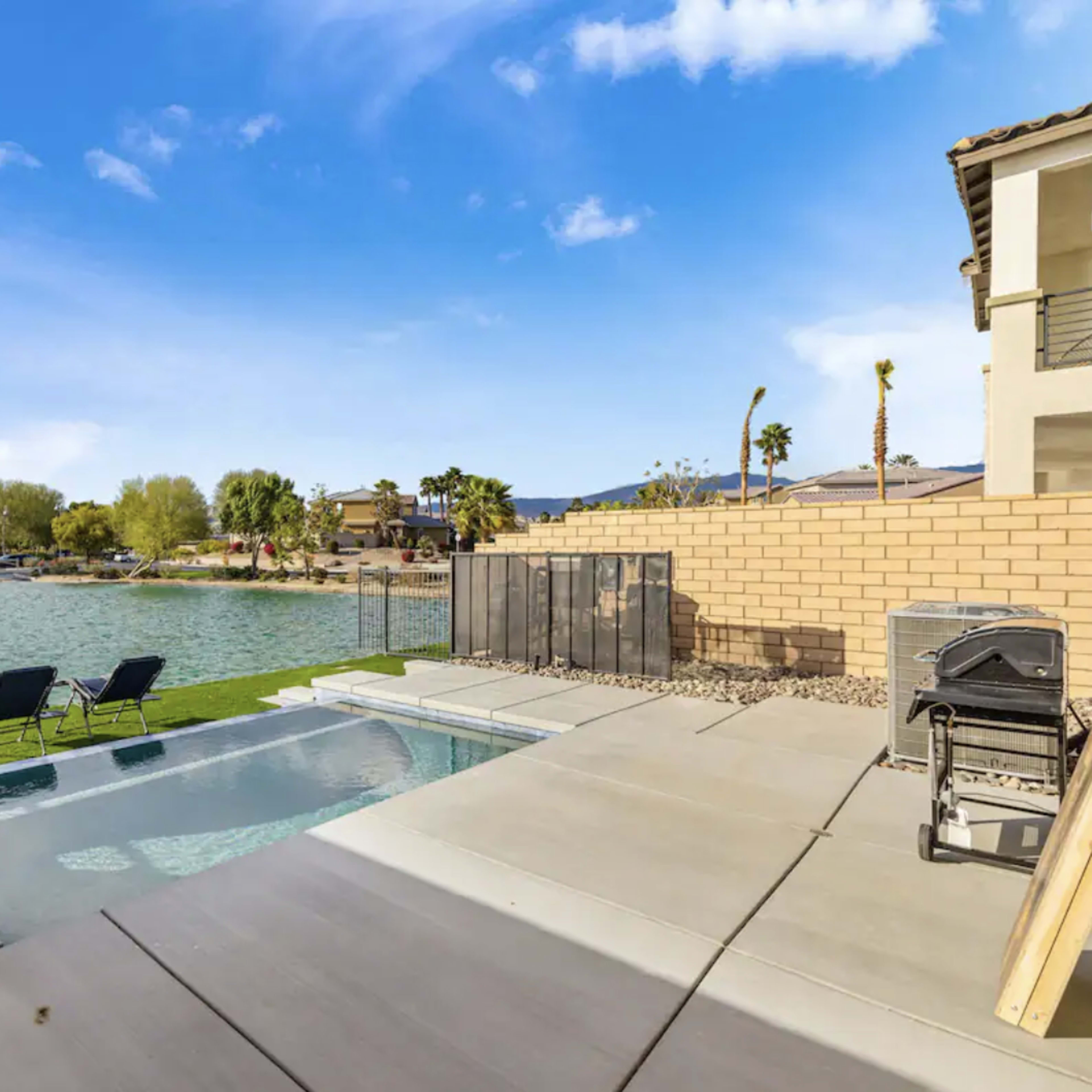 A backyard with a small pool, two lounge chairs, a grill, and a view of a lake and palm trees.