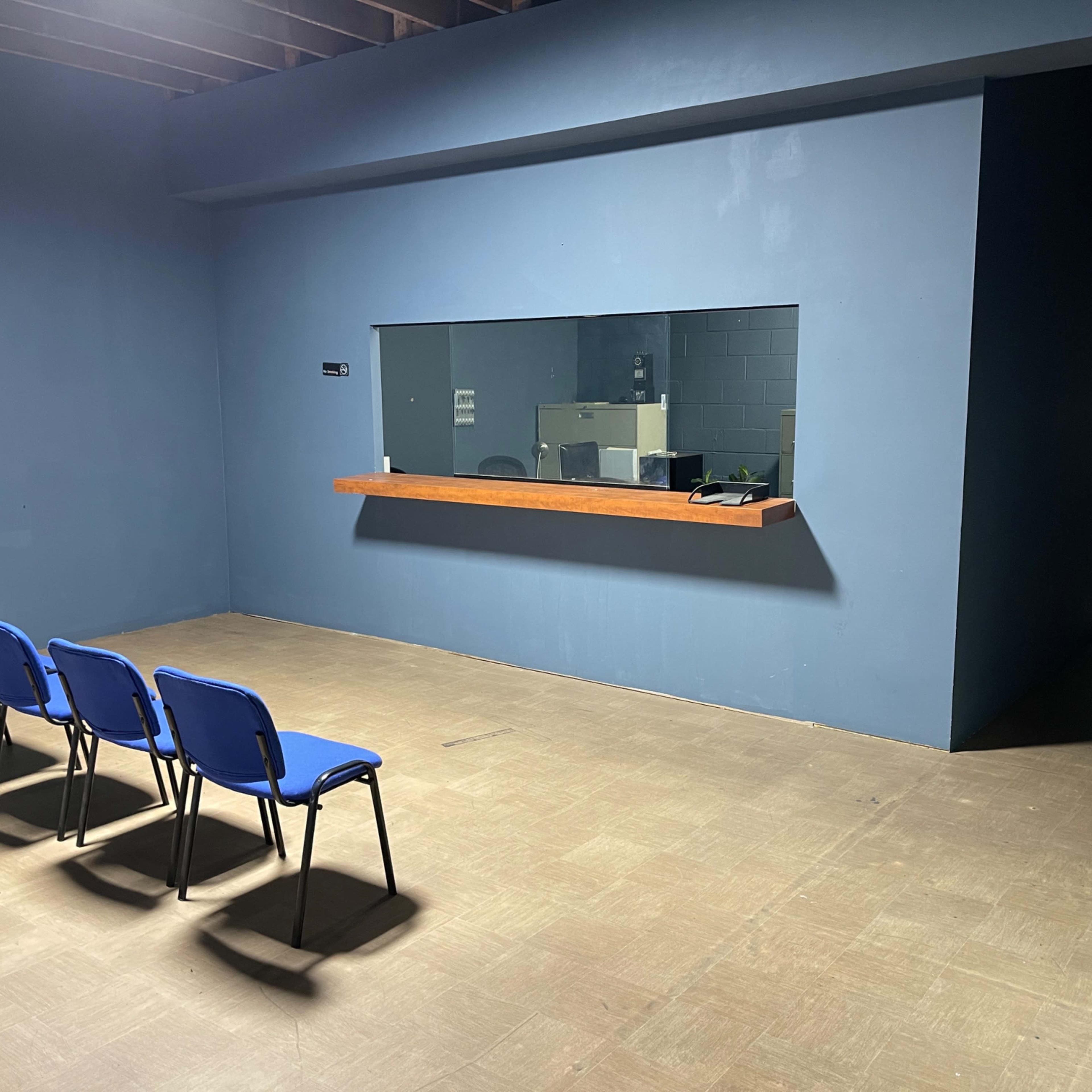 An empty room features four blue chairs arranged in a row facing a large window embedded in a blue wall.