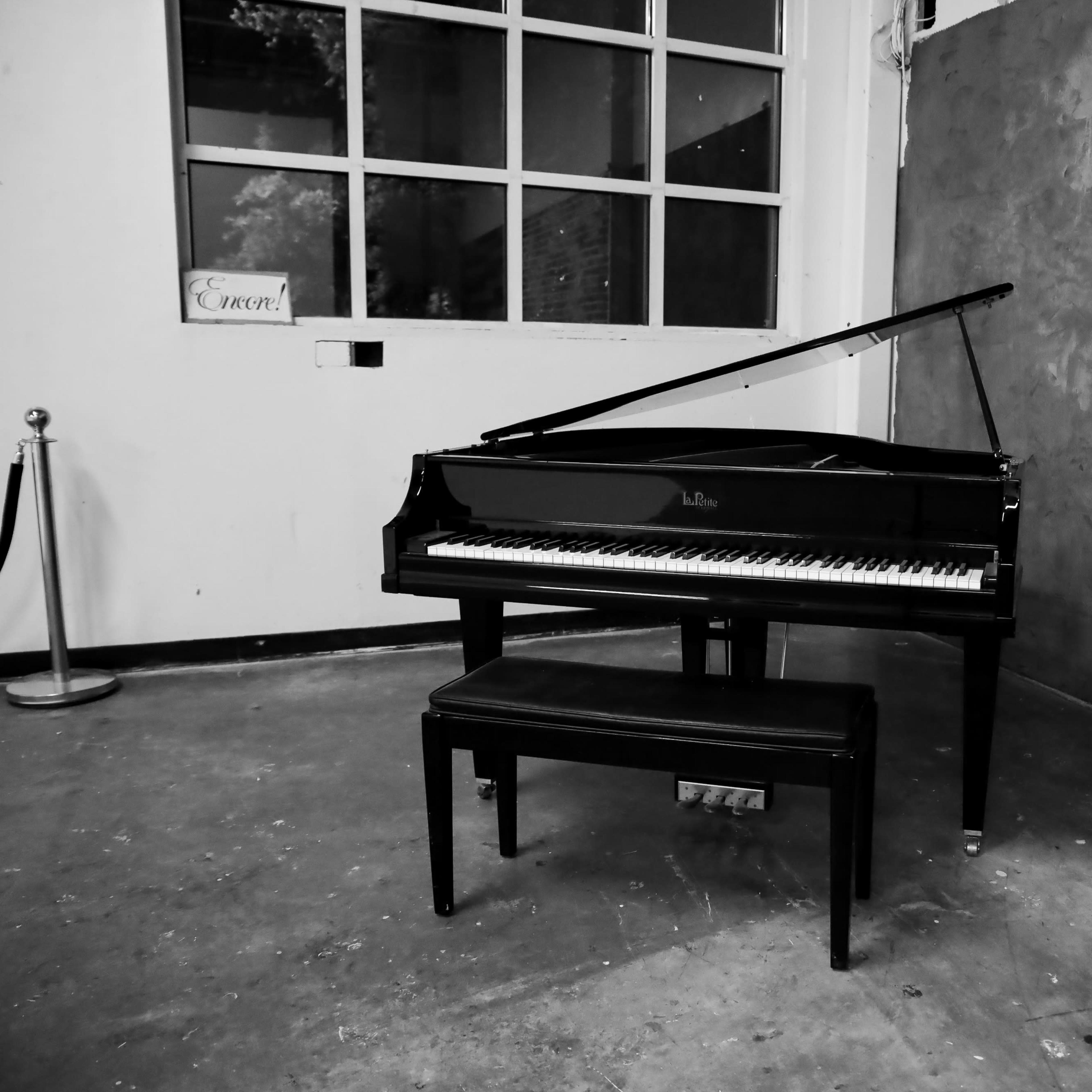A black grand piano and matching bench are positioned against a bare wall in a well-lit room.