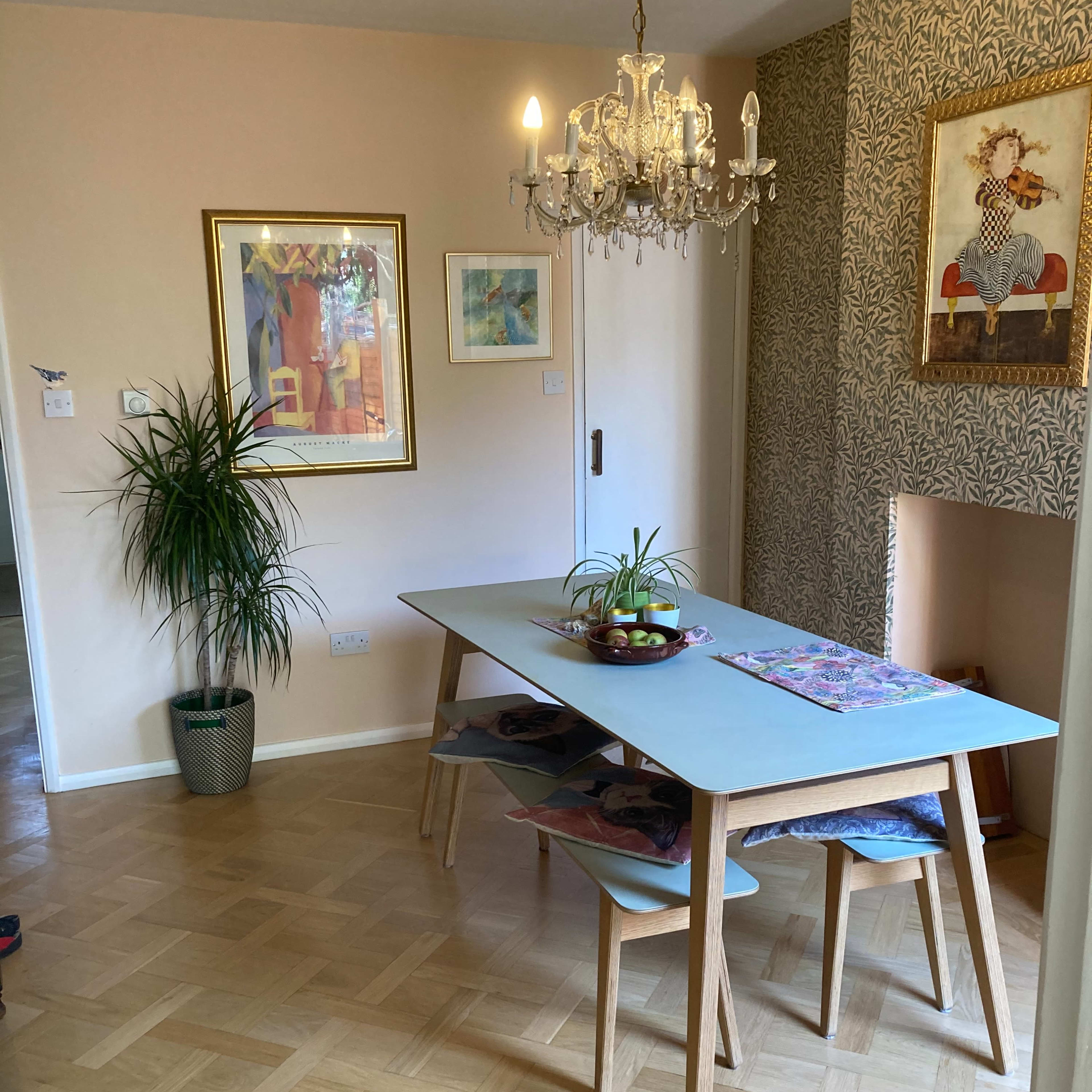 A light-filled dining area featuring a wooden table surrounded by chairs, a chandelier overhead, and wall decor including framed artwork and patterned wallpaper.