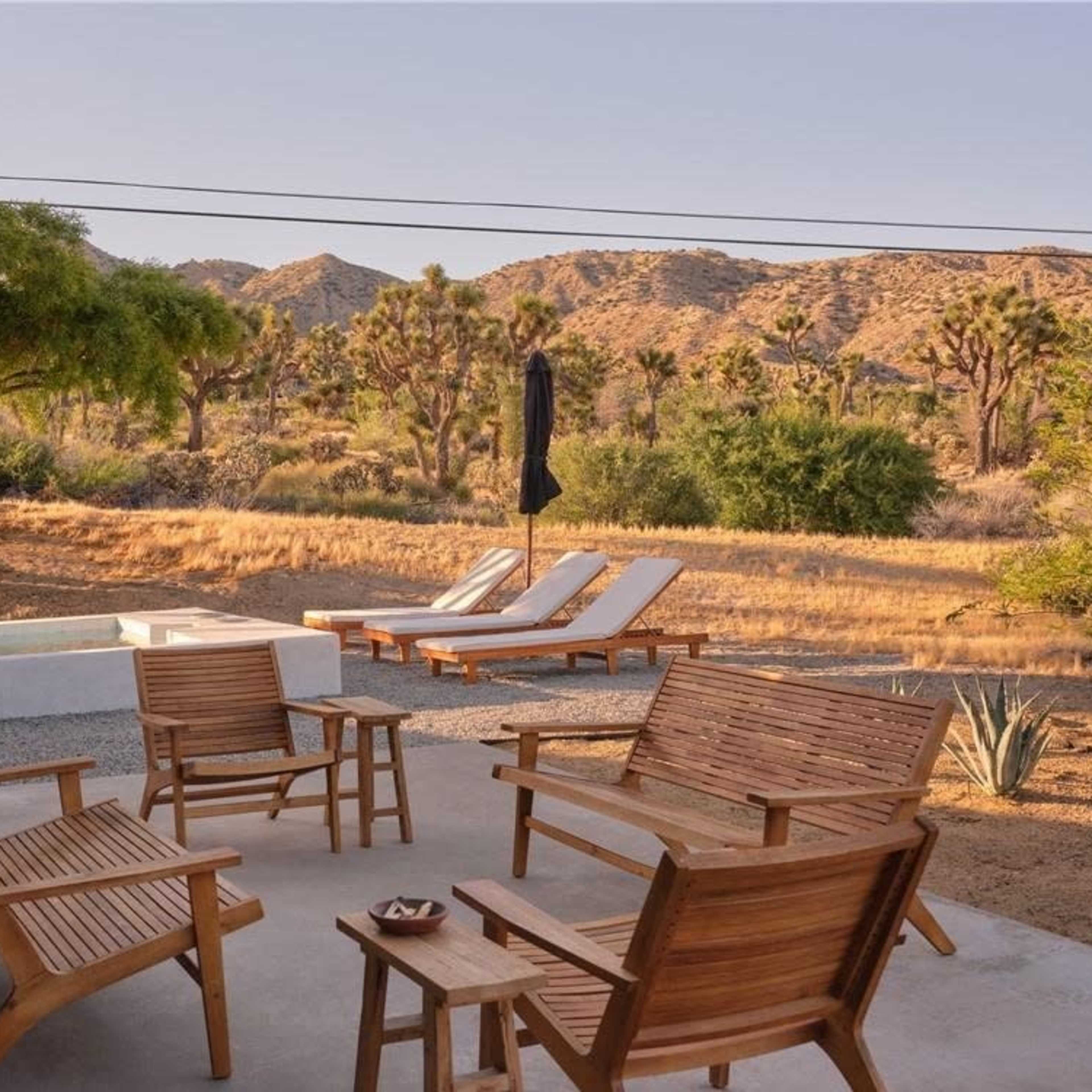 A wooden patio set with chairs surrounds a small table, overlooking a desert landscape with Joshua trees and mountains in the background.