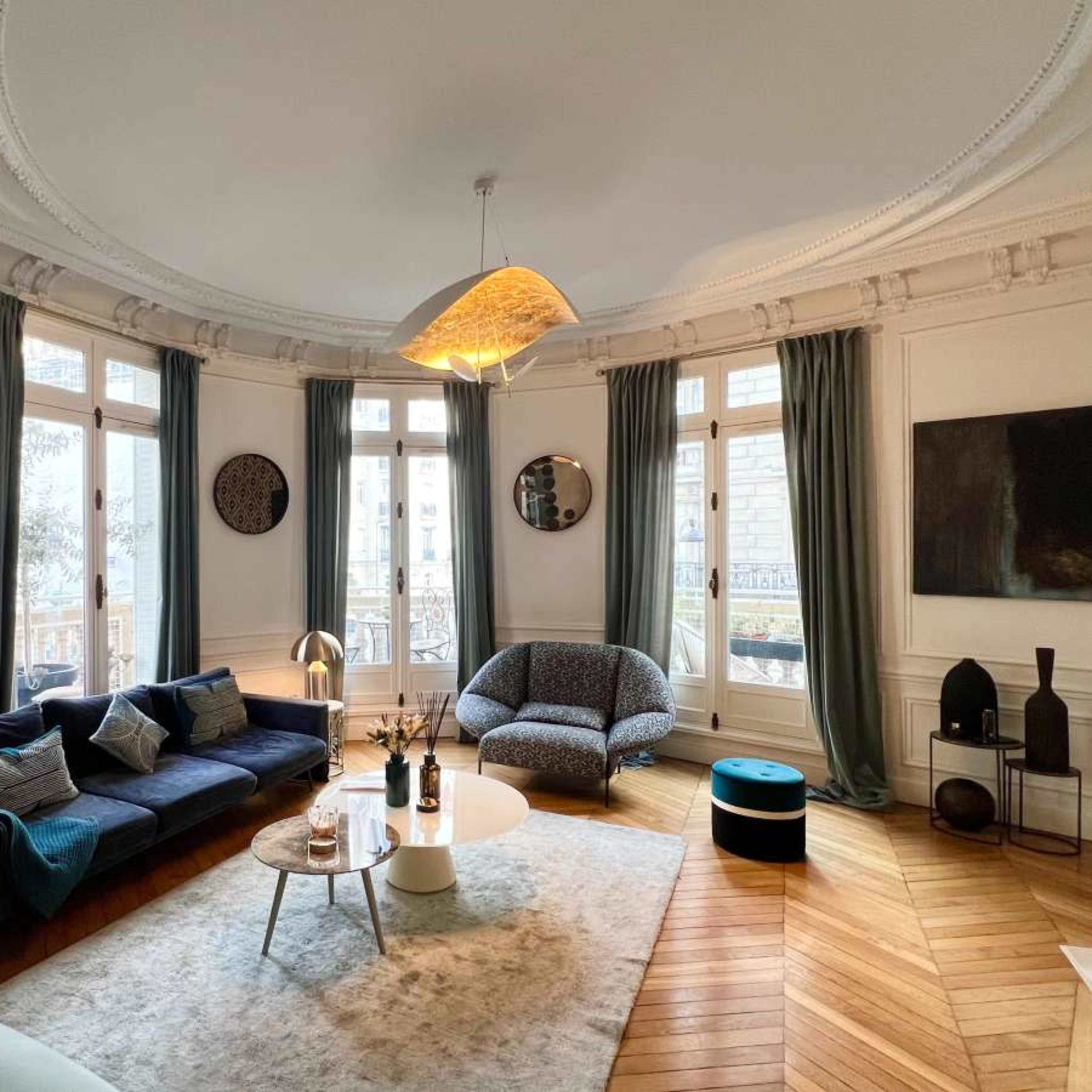 A furnished living room features a blue sofa, a circular coffee table, and large windows with curtains, all set on a patterned wooden floor.