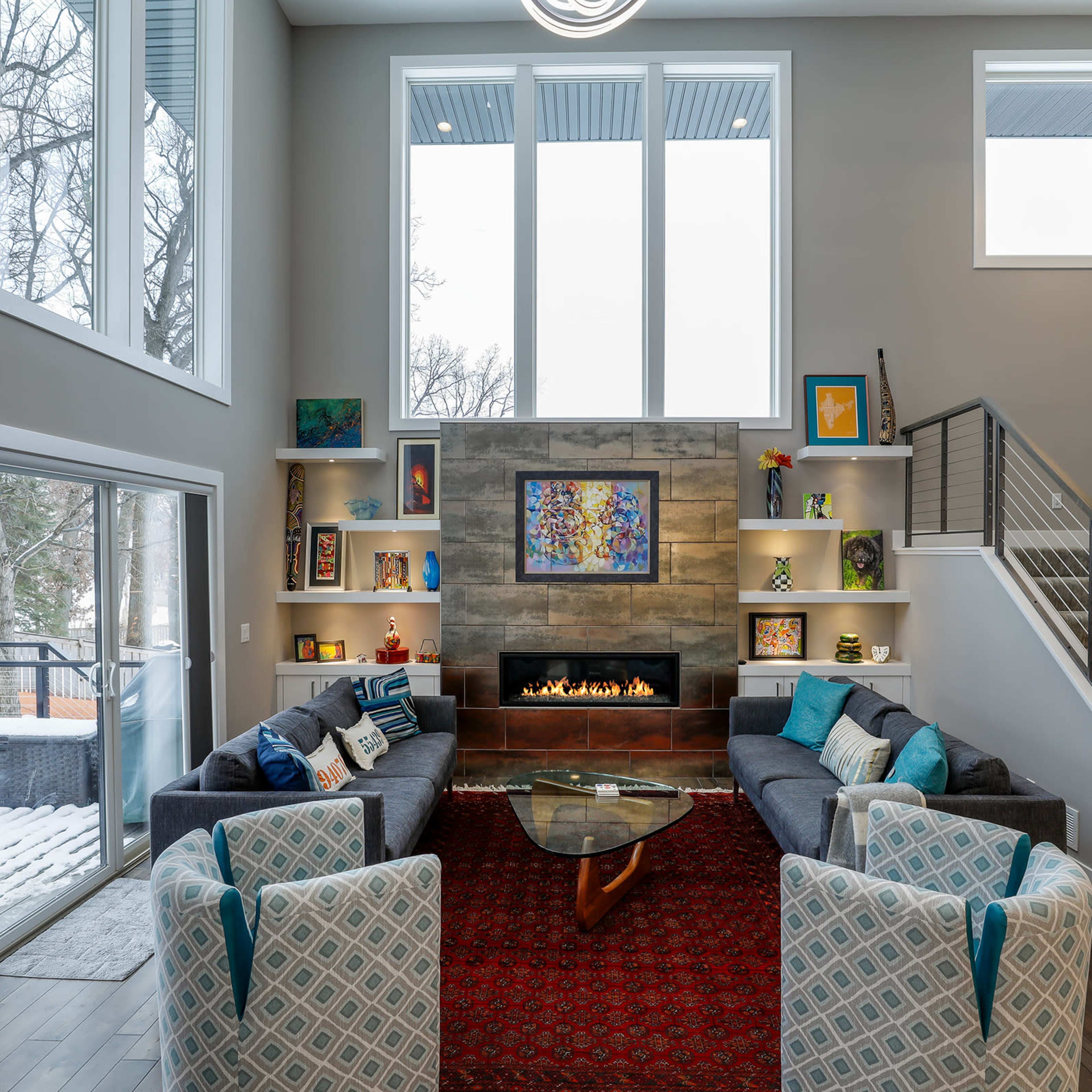 A modern living room with large windows, a central fireplace, an area rug, and a staircase leading to an upper level.