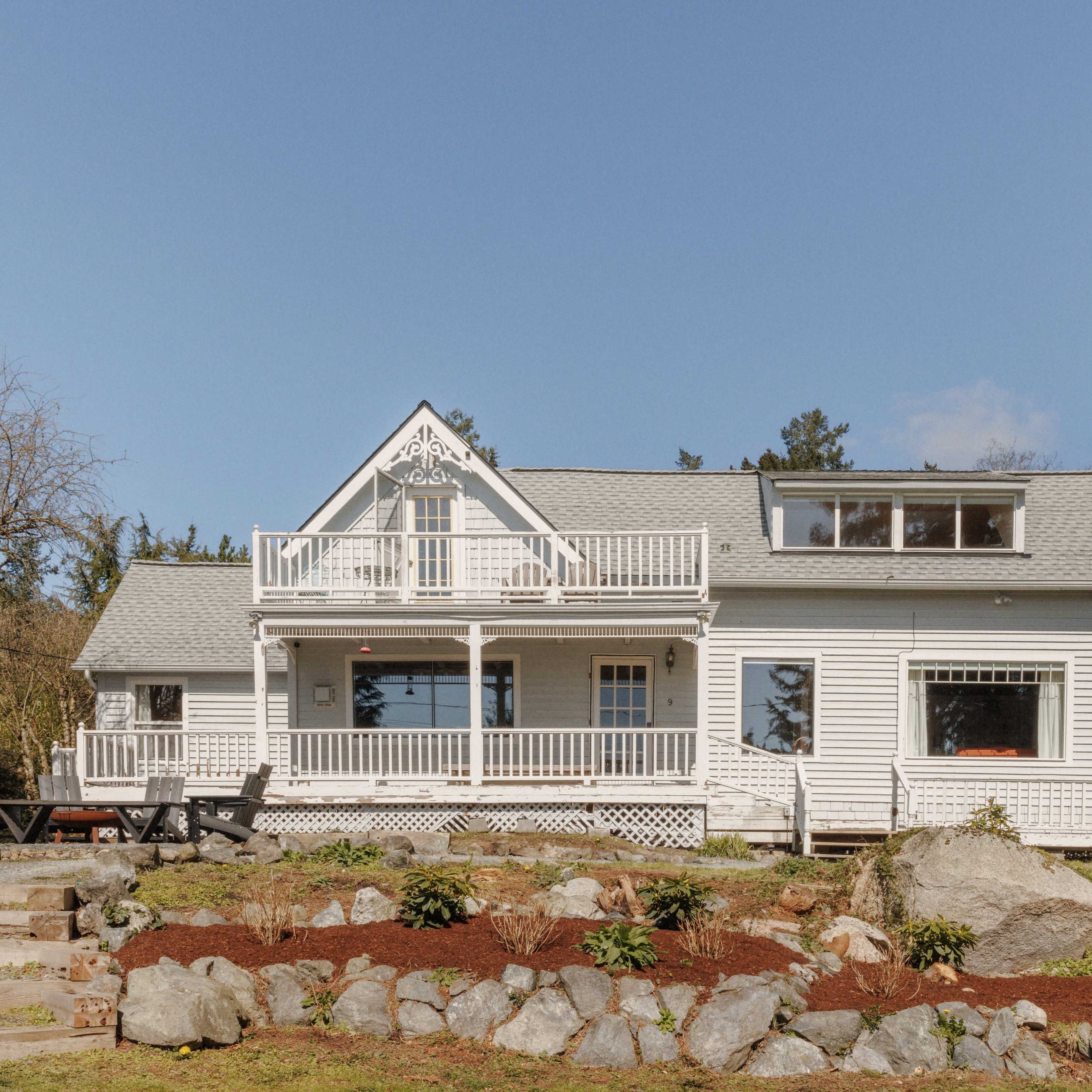 A large, white, two-story house with a front porch and balcony is surrounded by a landscaped yard featuring rocks and greenery.