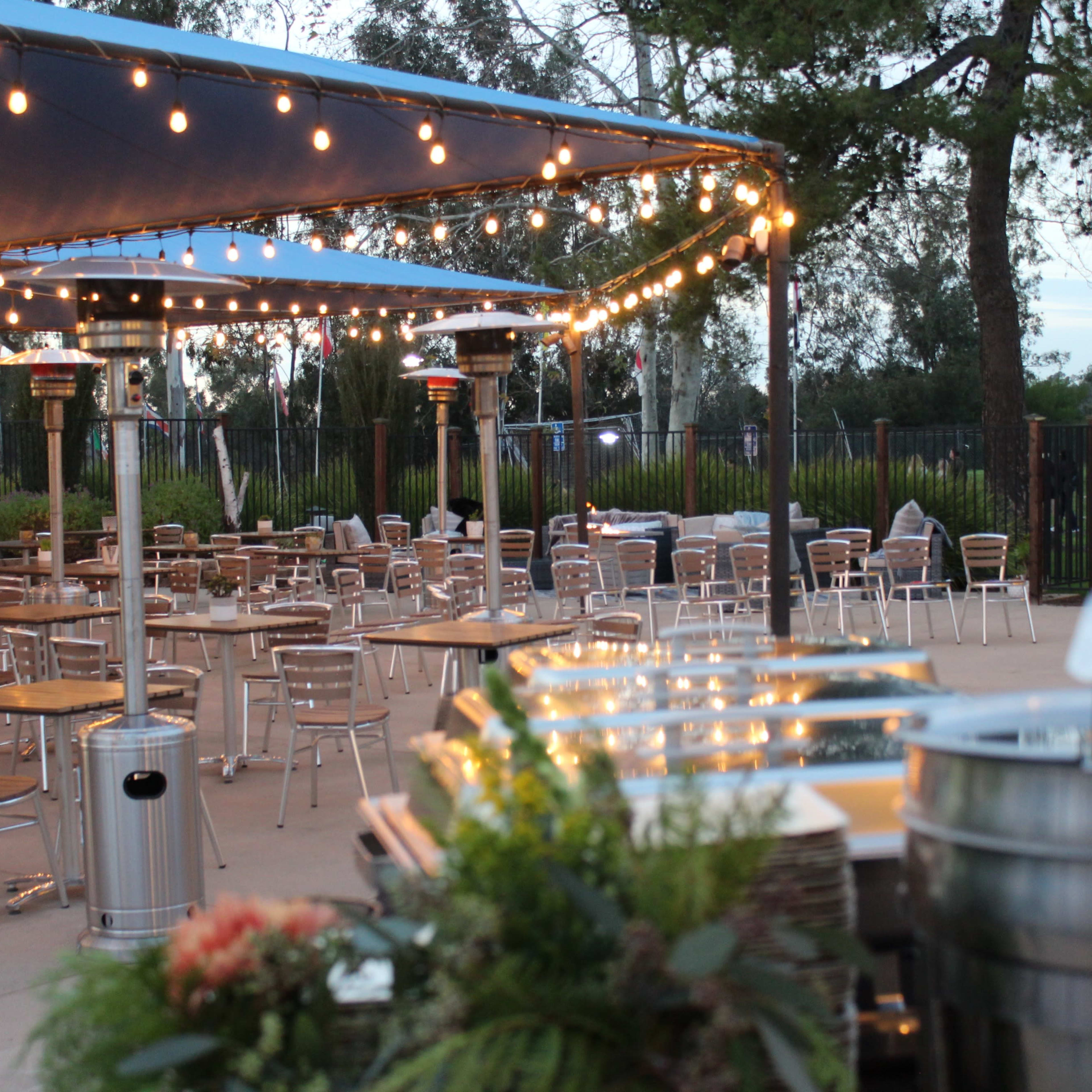 An outdoor dining area features tables and chairs under a large tent with string lights and heating lamps, set against a landscaped background.