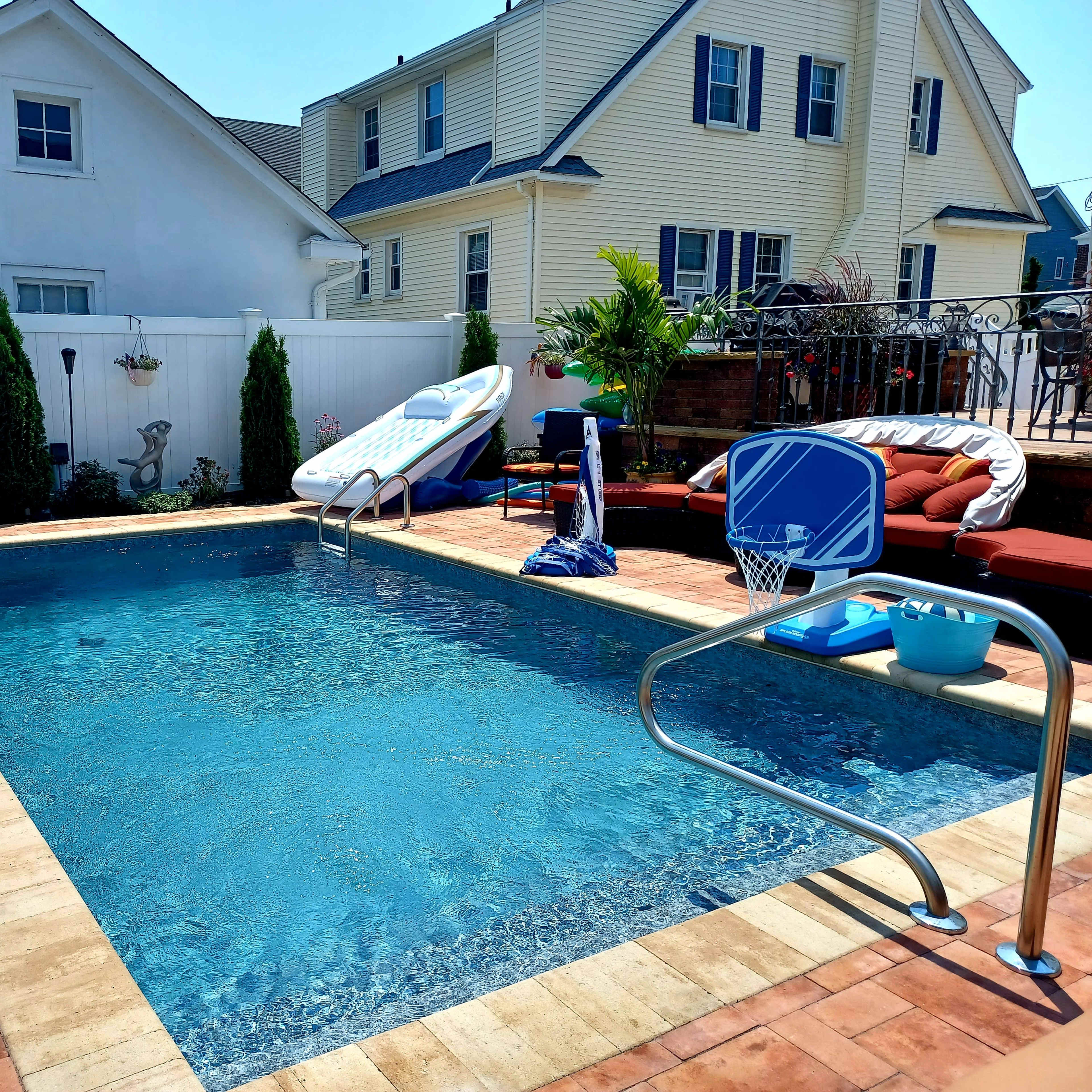 The scene depicts a backyard swimming pool surrounded by a deck, a slide, lounge furniture, and a basketball hoop.