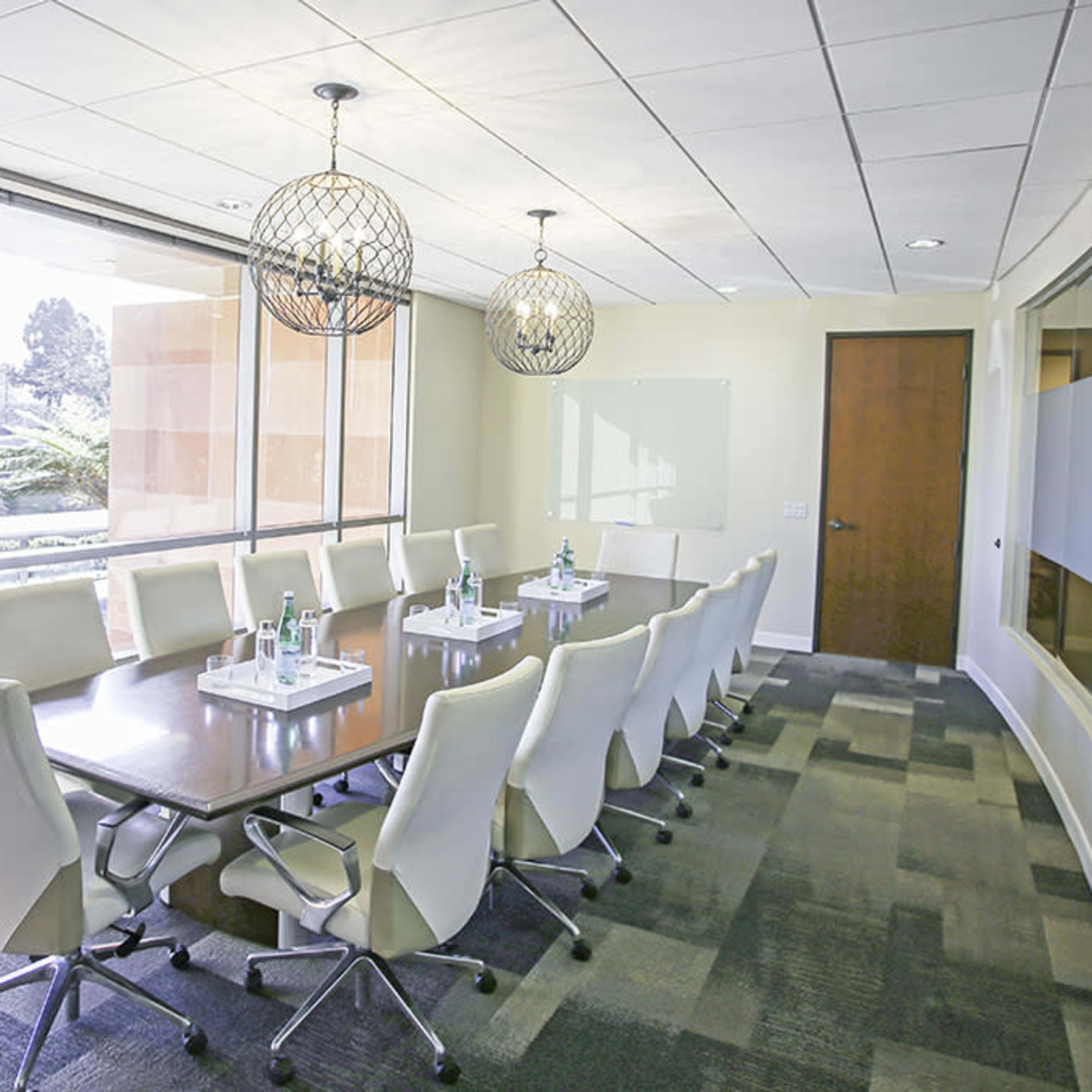 A modern conference room with a long table surrounded by chairs, large windows allowing natural light, and decorative light fixtures hanging above.