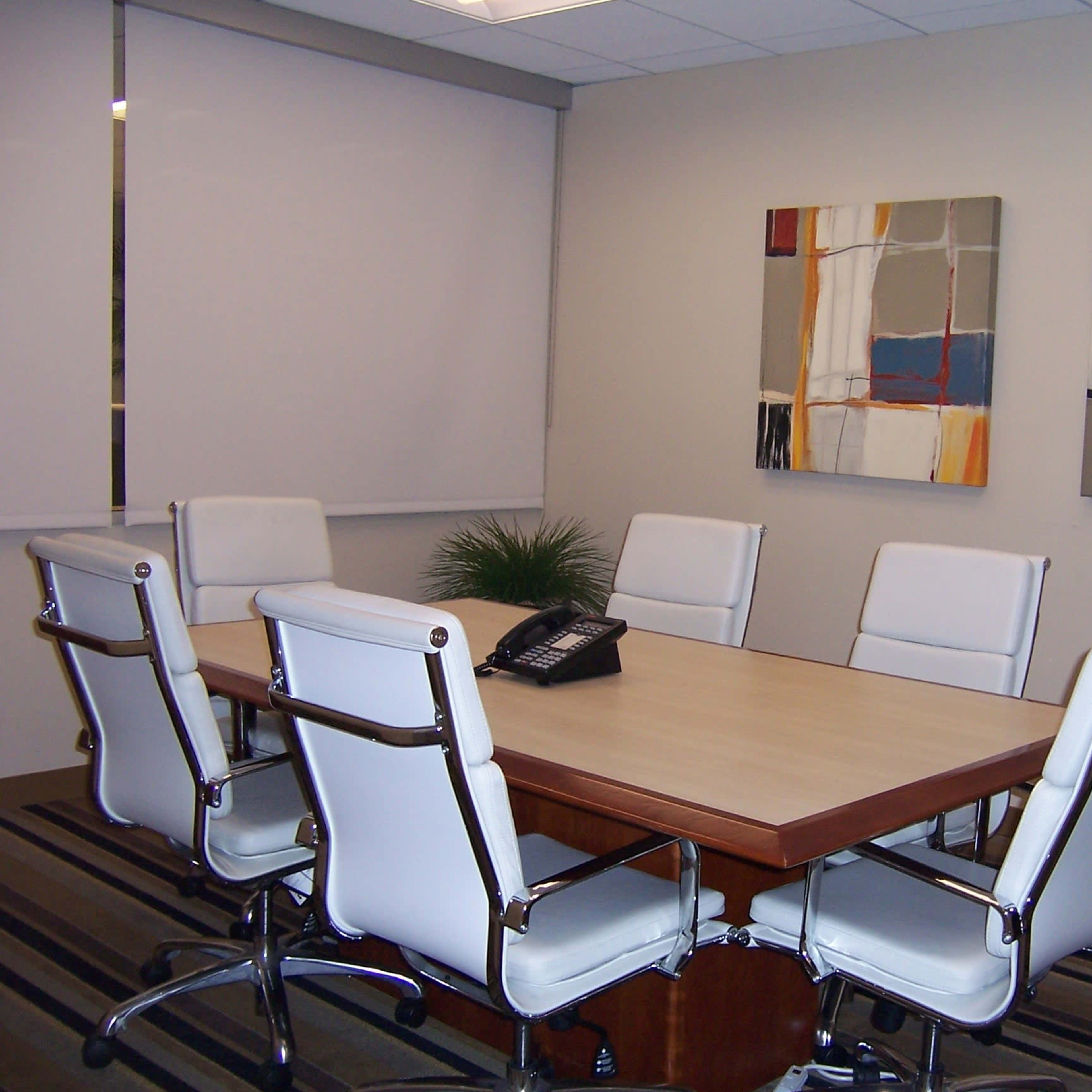 A conference room is set up with a rectangular table surrounded by six white chairs, and two abstract paintings are mounted on the wall.
