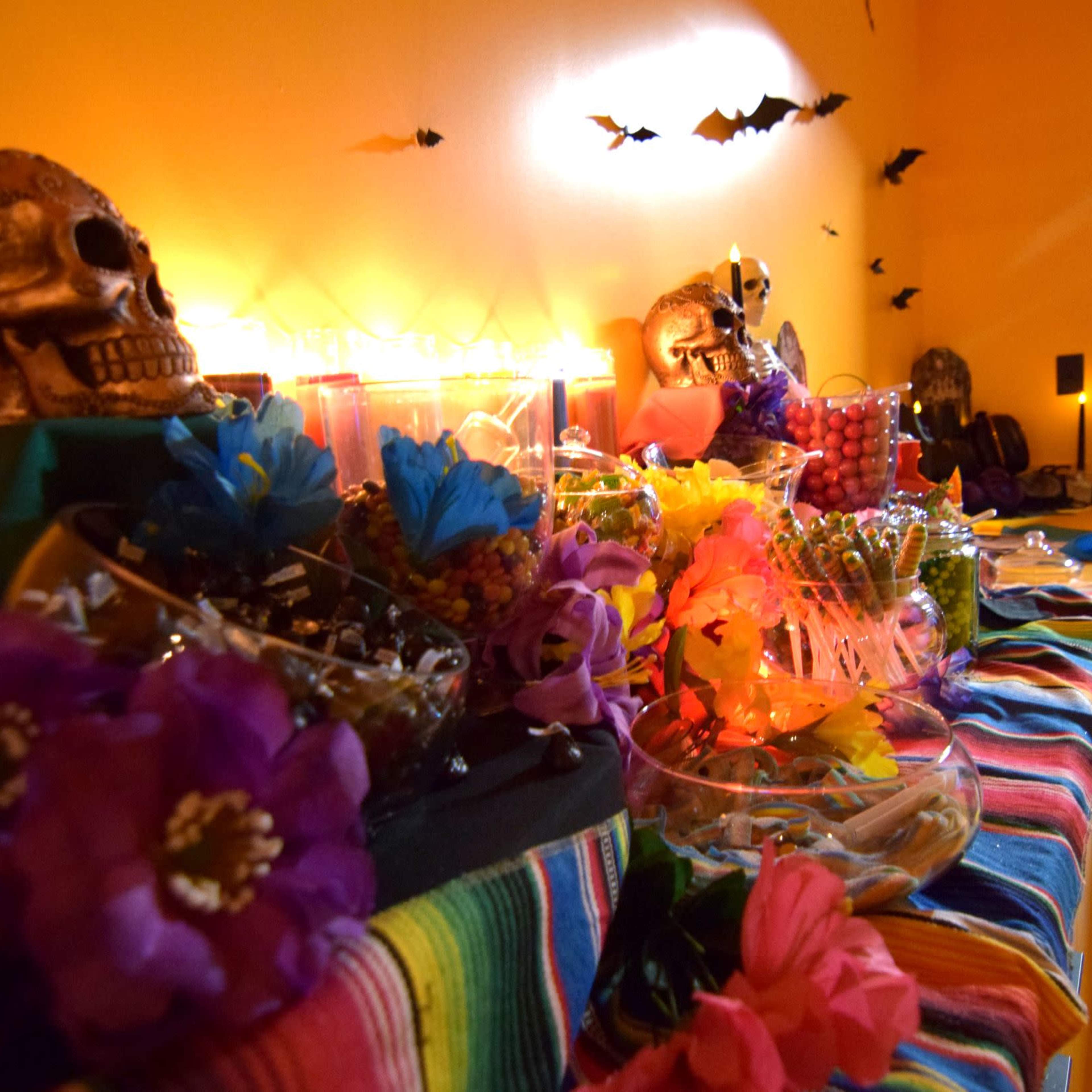A decorated table features colorful candies, flowers, and skull decorations against a bright yellow wall.