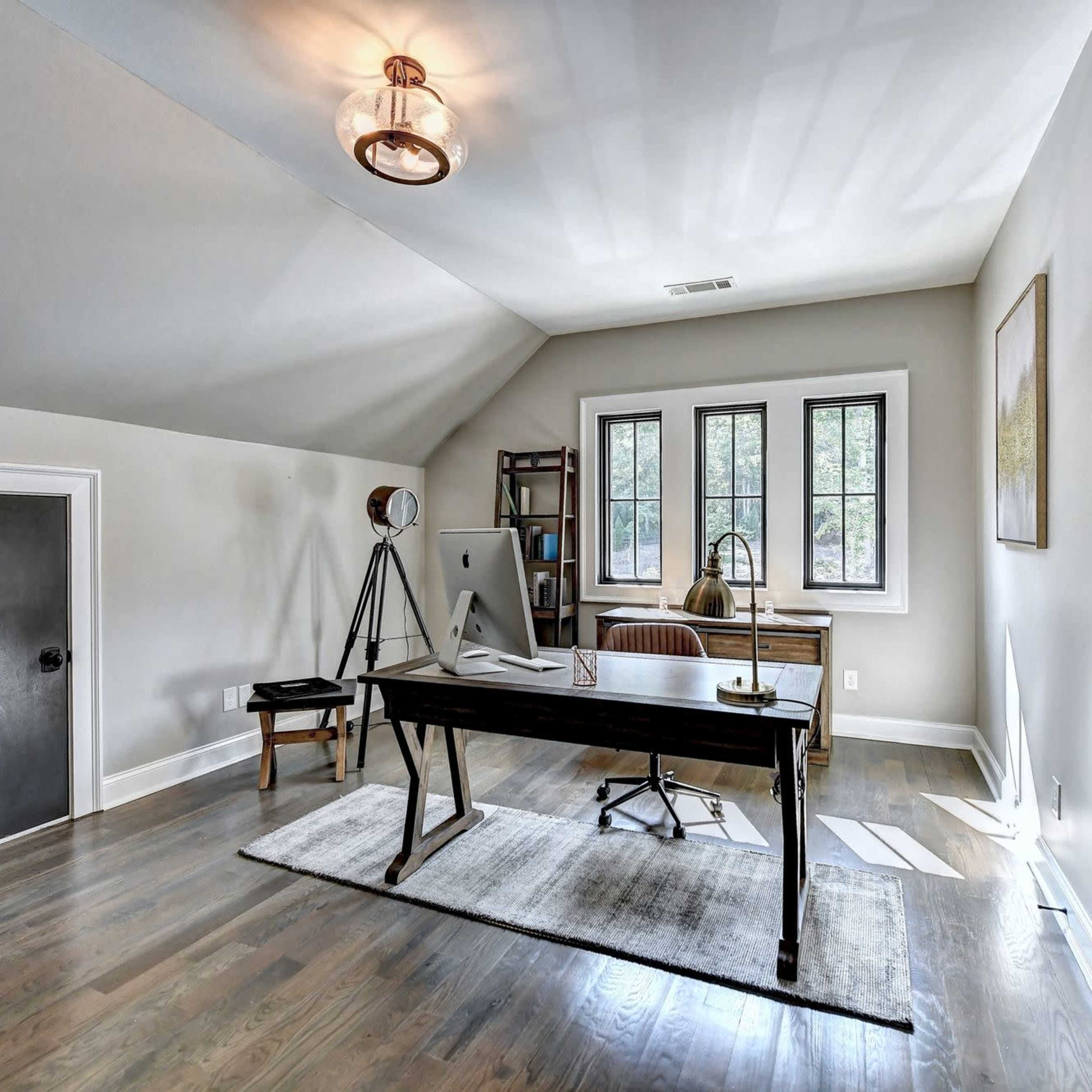 A bright, modern home office with a wooden desk, a chair, and large windows, complemented by a bookshelf and a tripod lamp.