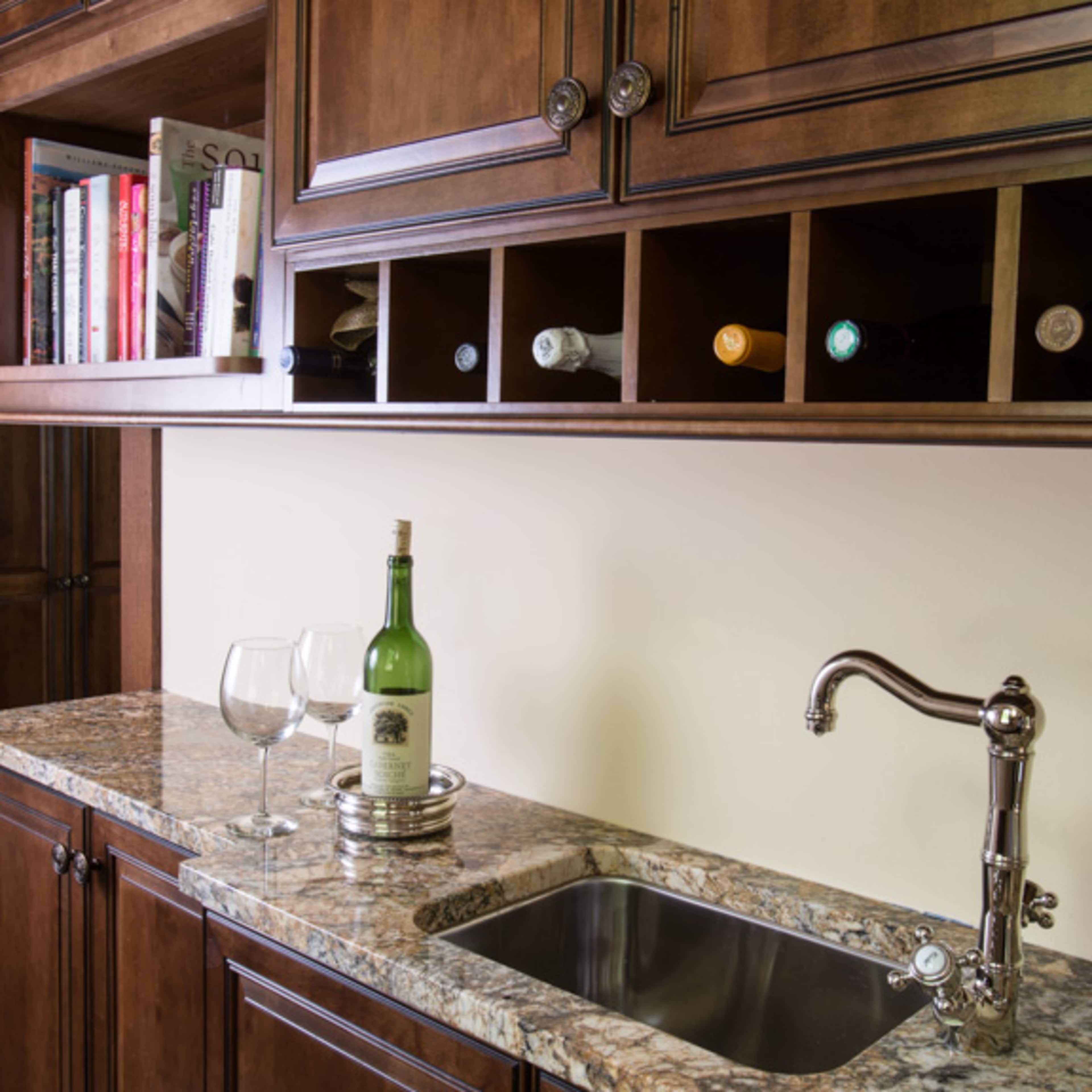 A kitchen area with a granite countertop, a sink, a wine bottle, and two glasses, along with wooden cabinets and a shelf holding several bottles.