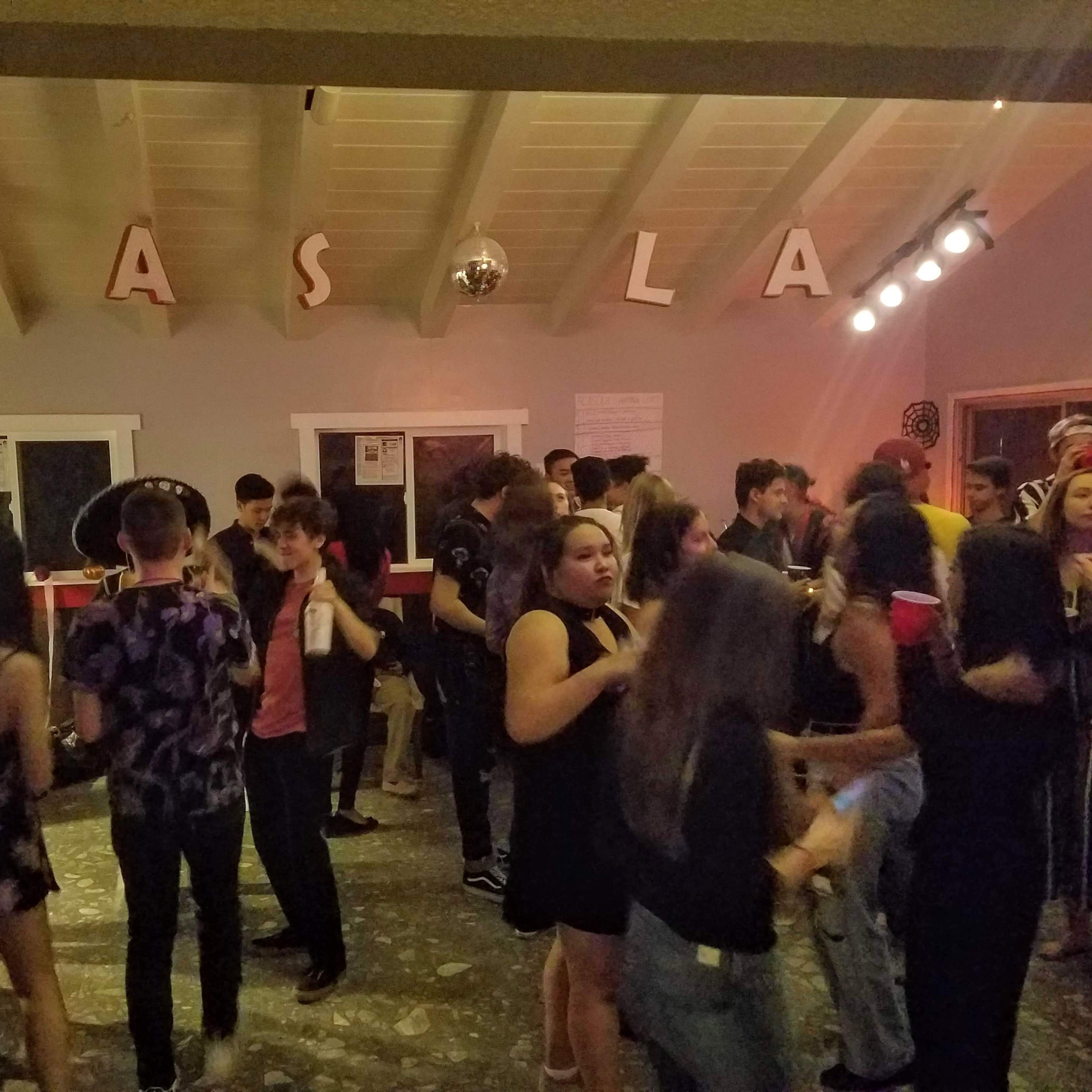 A crowd of people socializes and dances in a dimly lit room decorated with the word "CASA" on the wall.