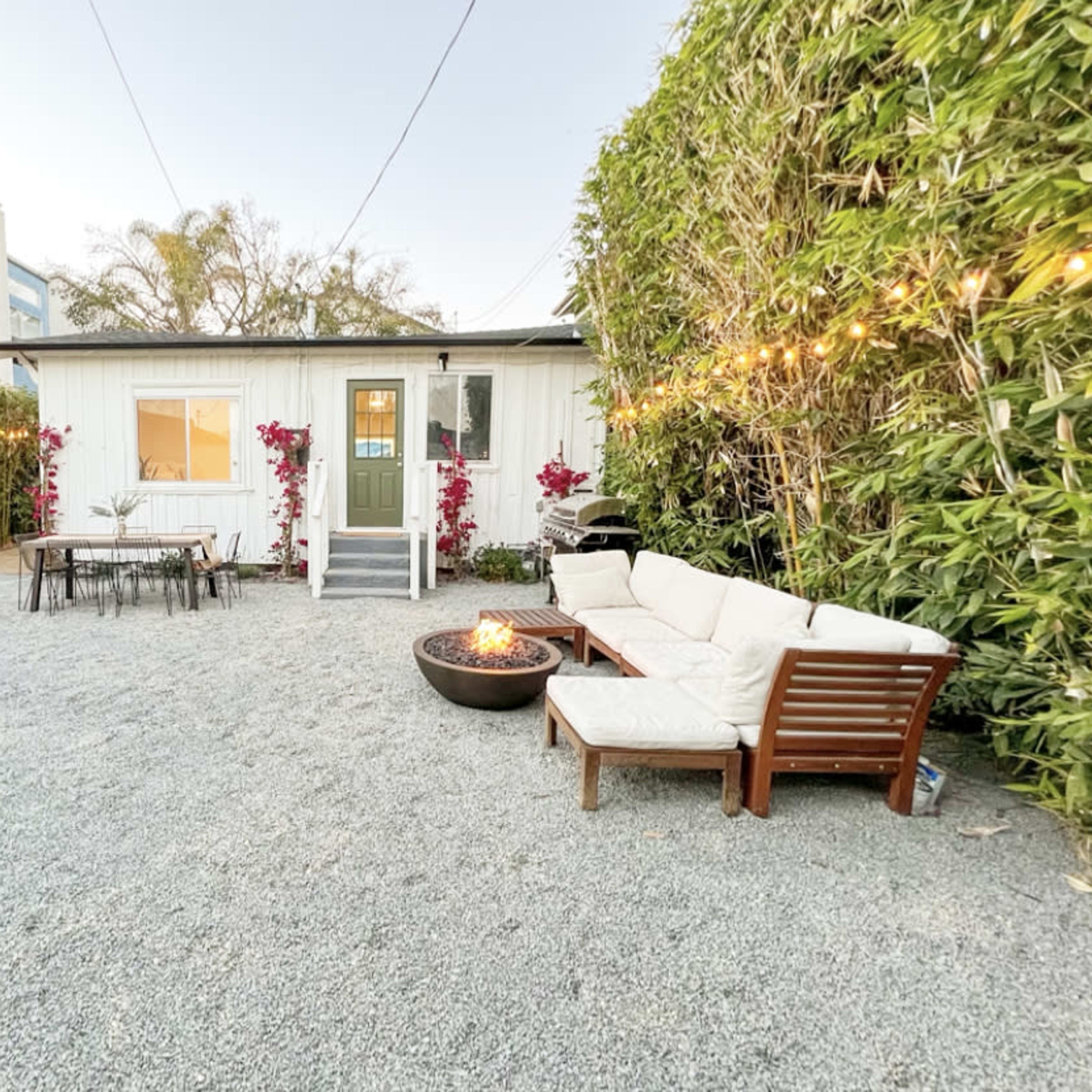 A small white cottage is surrounded by bamboo and gravel, with outdoor seating and string lights illuminating the area in the evening.