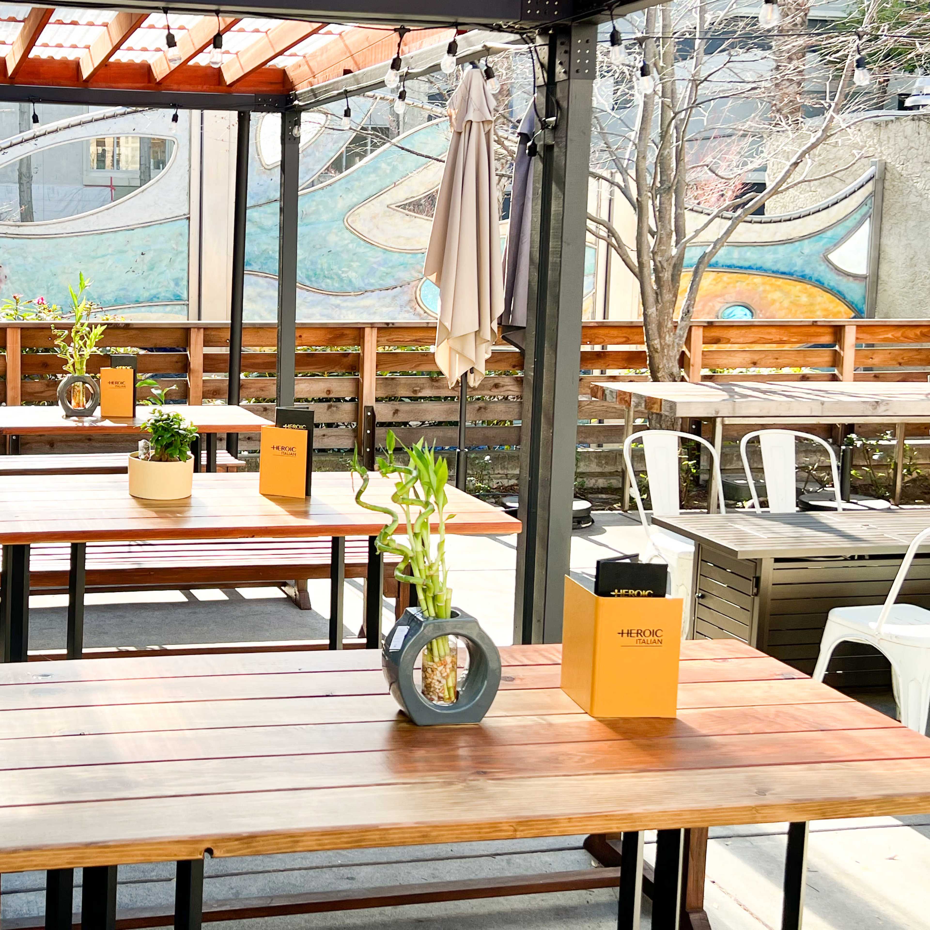 An outdoor dining area features wooden tables with potted plants and a colorful mural in the background.