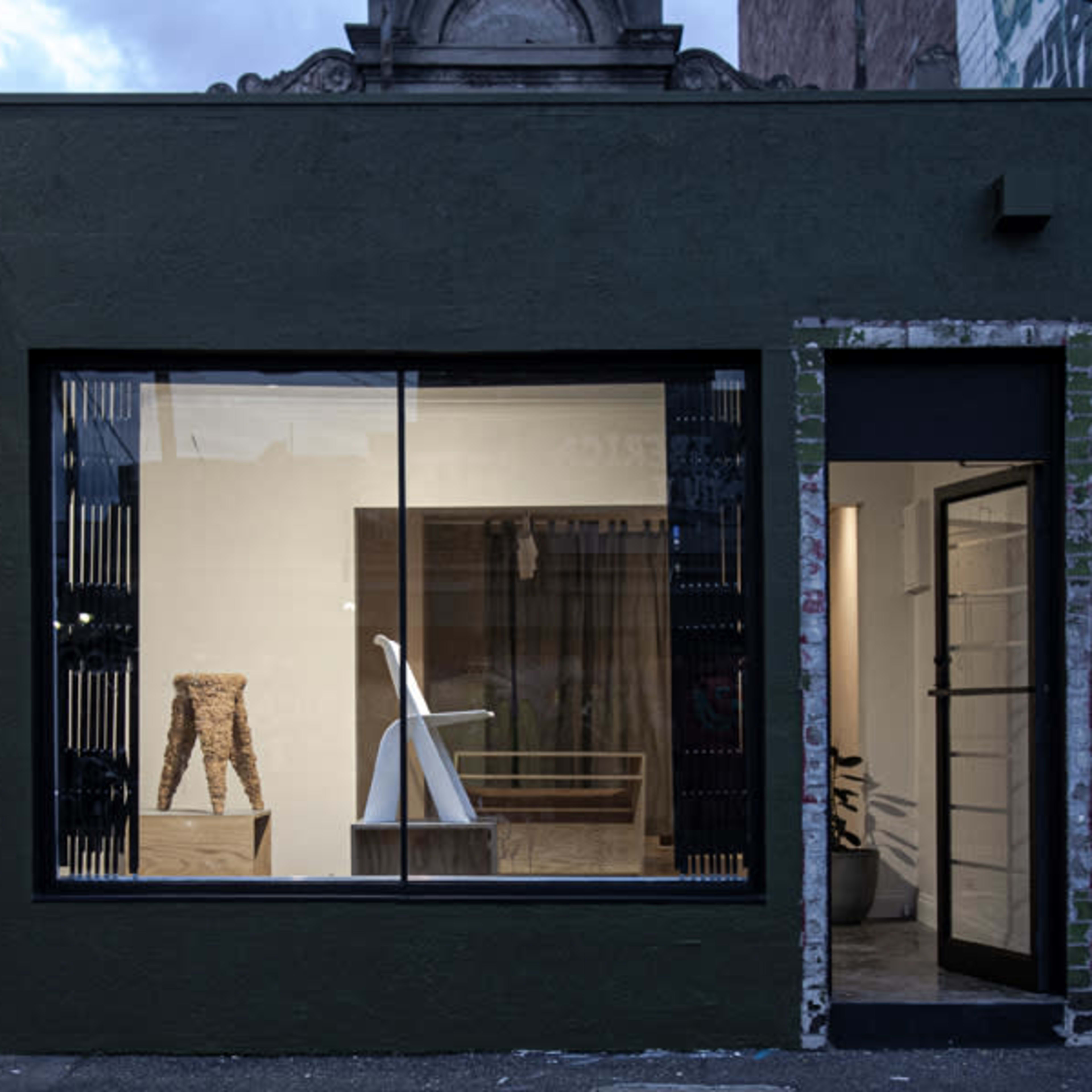 The exterior of a modern dark green building with large windows displaying minimalist furniture and an interior featuring a clean, open space with a wooden table and metal shelving.