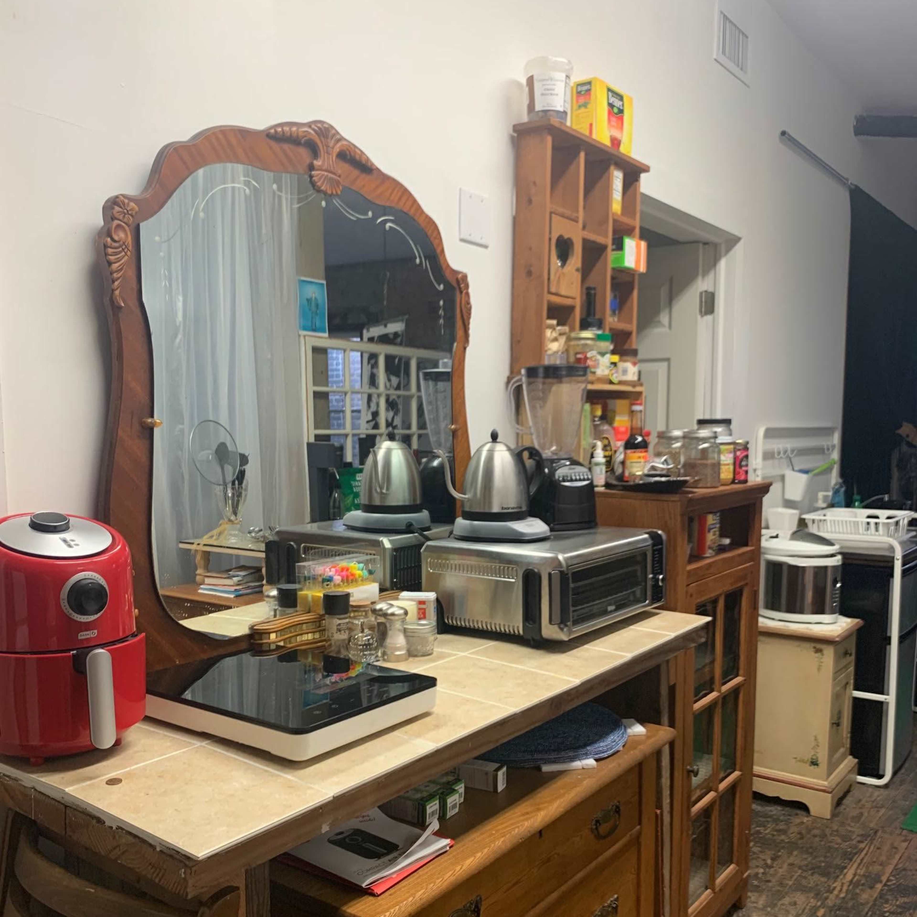 A wooden table displays various kitchen appliances and a large mirror, alongside a tall cabinet and additional storage.