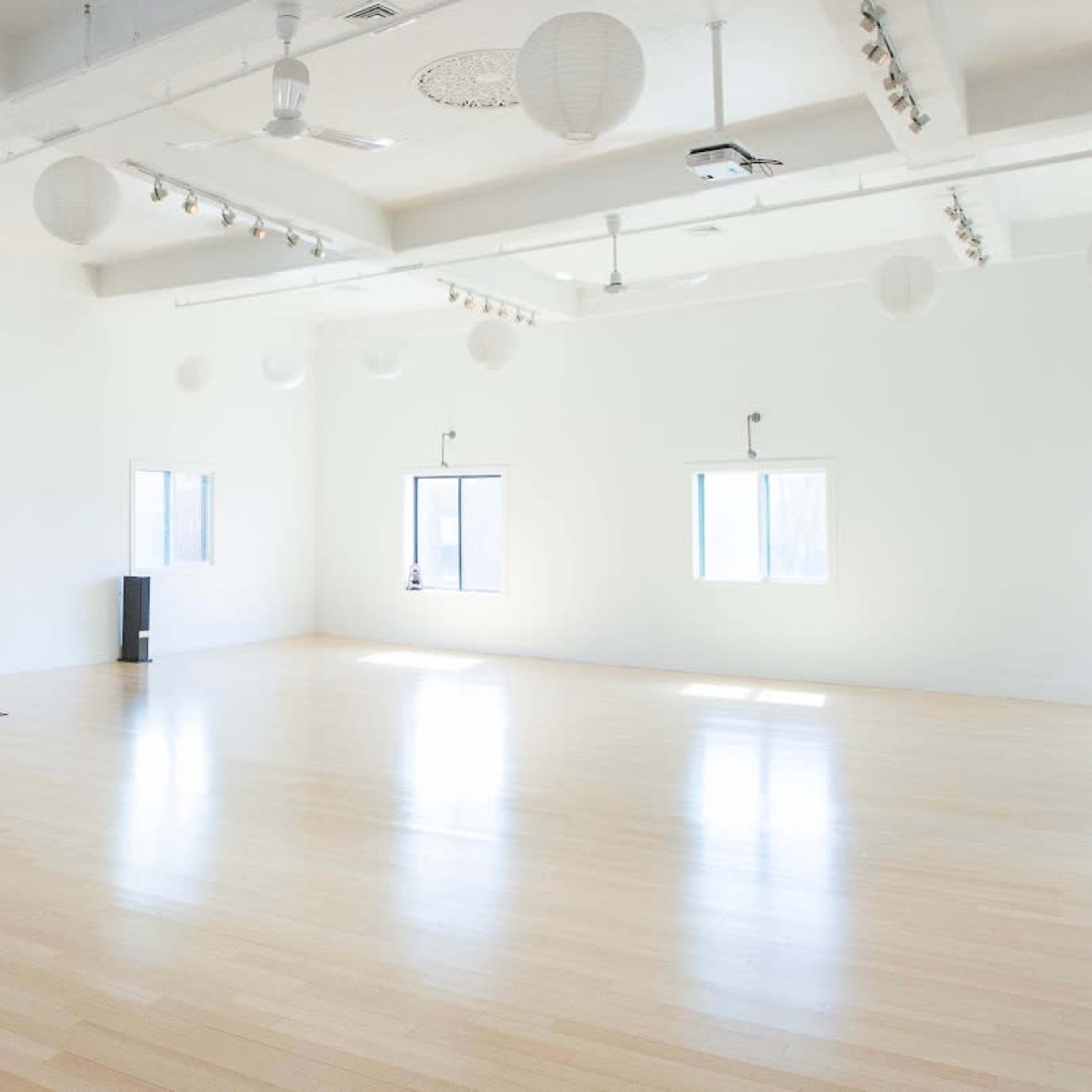 A spacious, empty studio with wooden flooring, three windows, and several hanging paper lanterns.