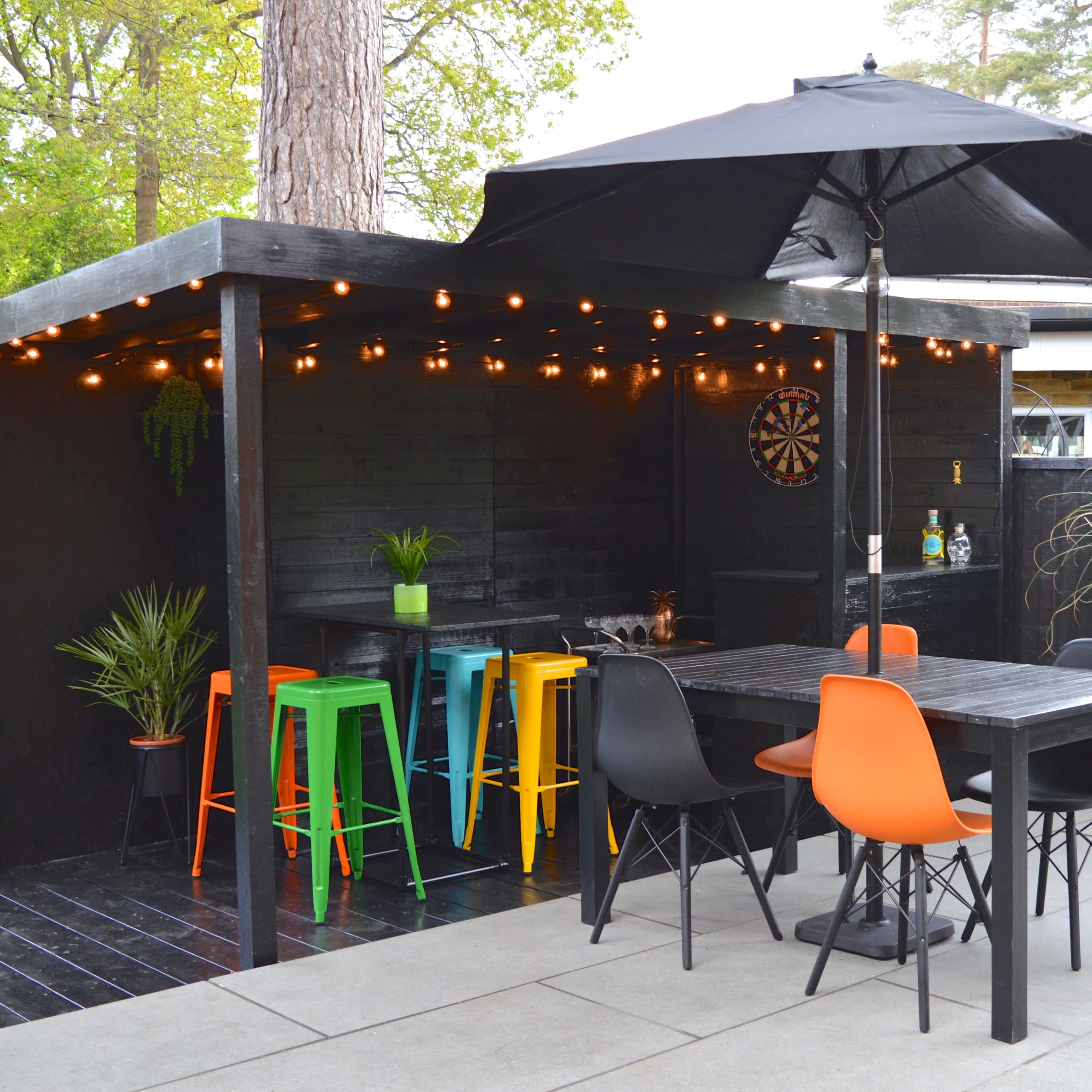 A black outdoor patio featuring colorful bar stools, a dining table, and string lights overhead in a garden setting.