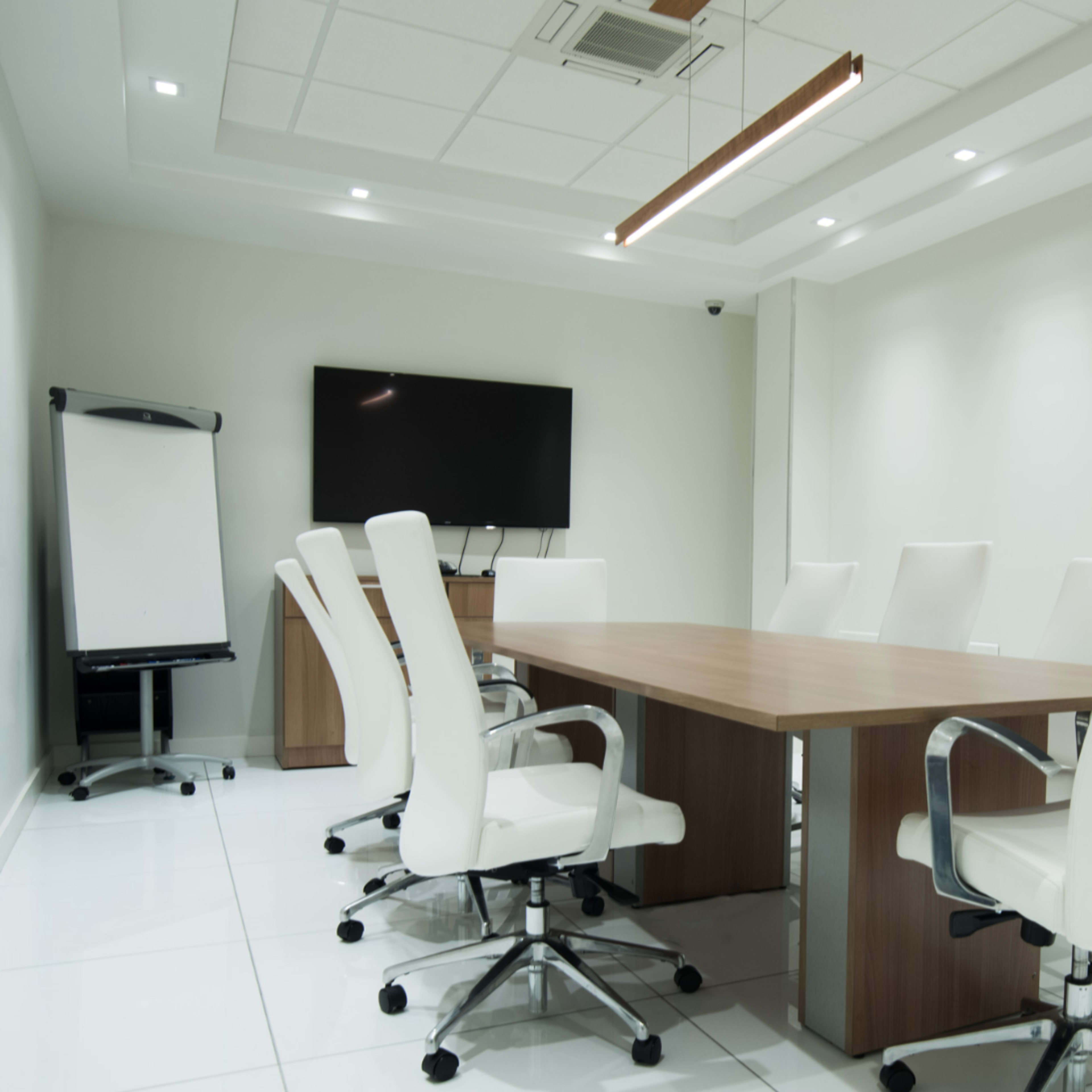 A modern conference room features a large wooden table surrounded by white ergonomic chairs, with a television screen and a flip chart on the walls.