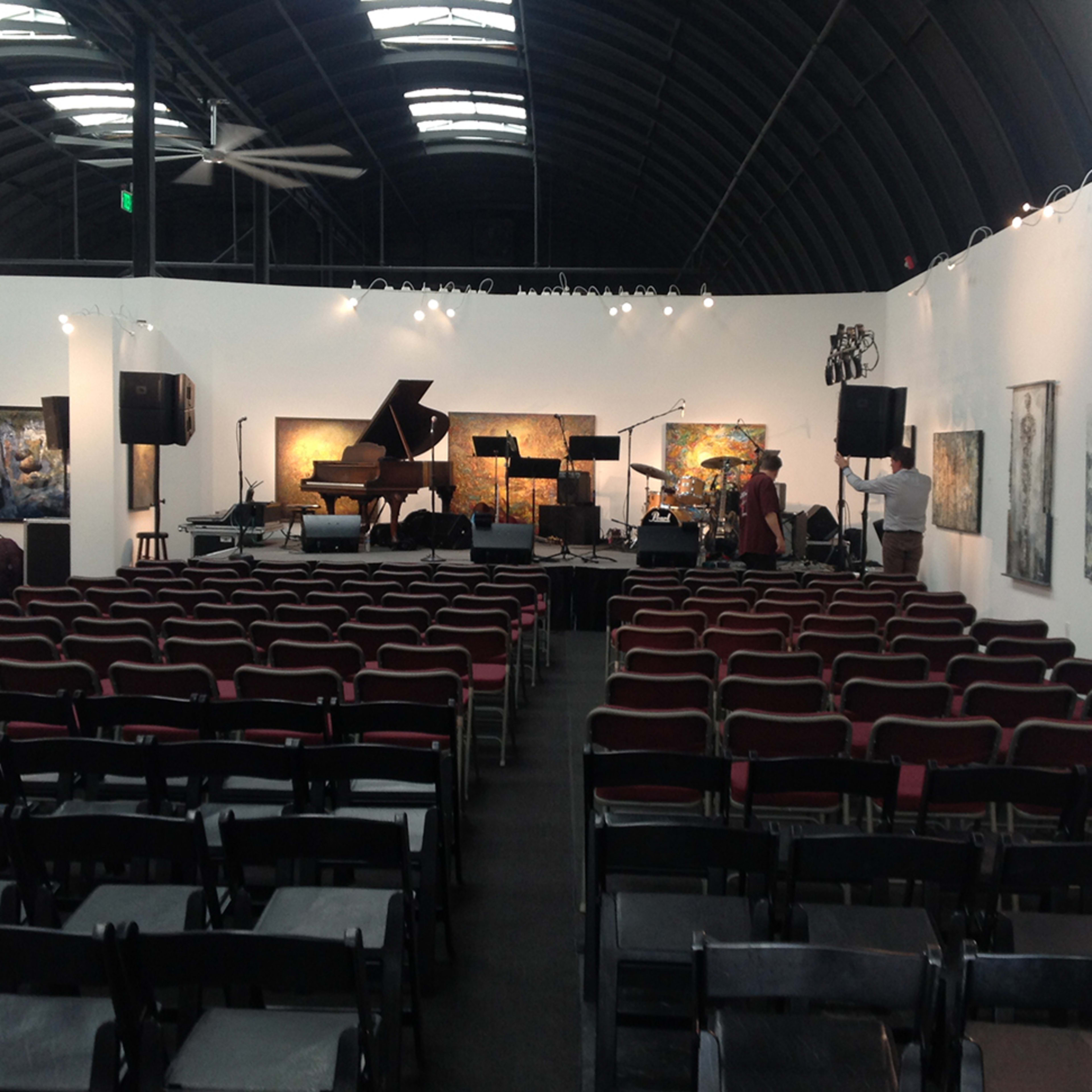 An empty performance space with rows of black chairs facing a grand piano and a backdrop of artwork on the walls.