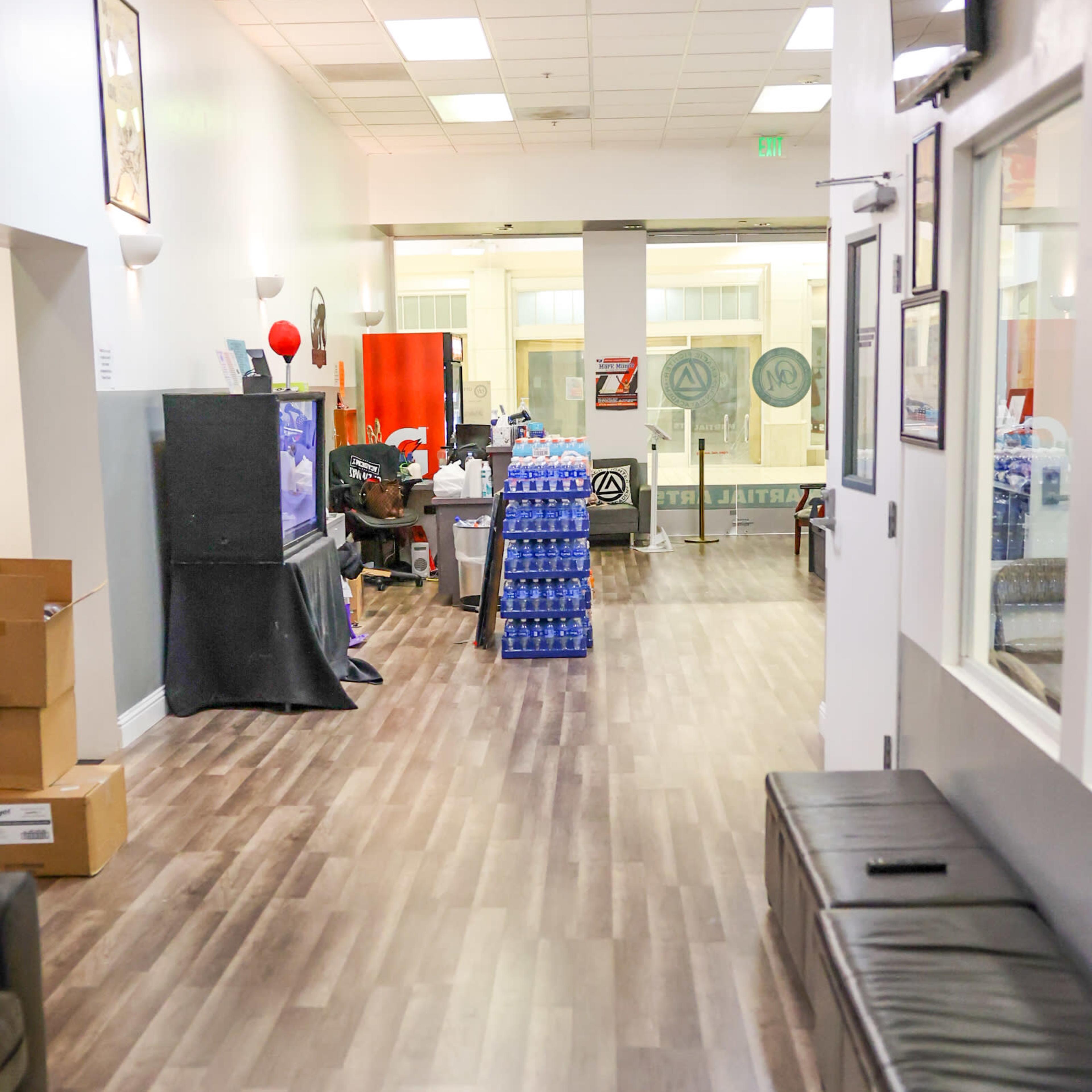 A long corridor with wooden flooring, lined with boxes and a stack of blue plastic water bottles, leading to a reception area in the background.