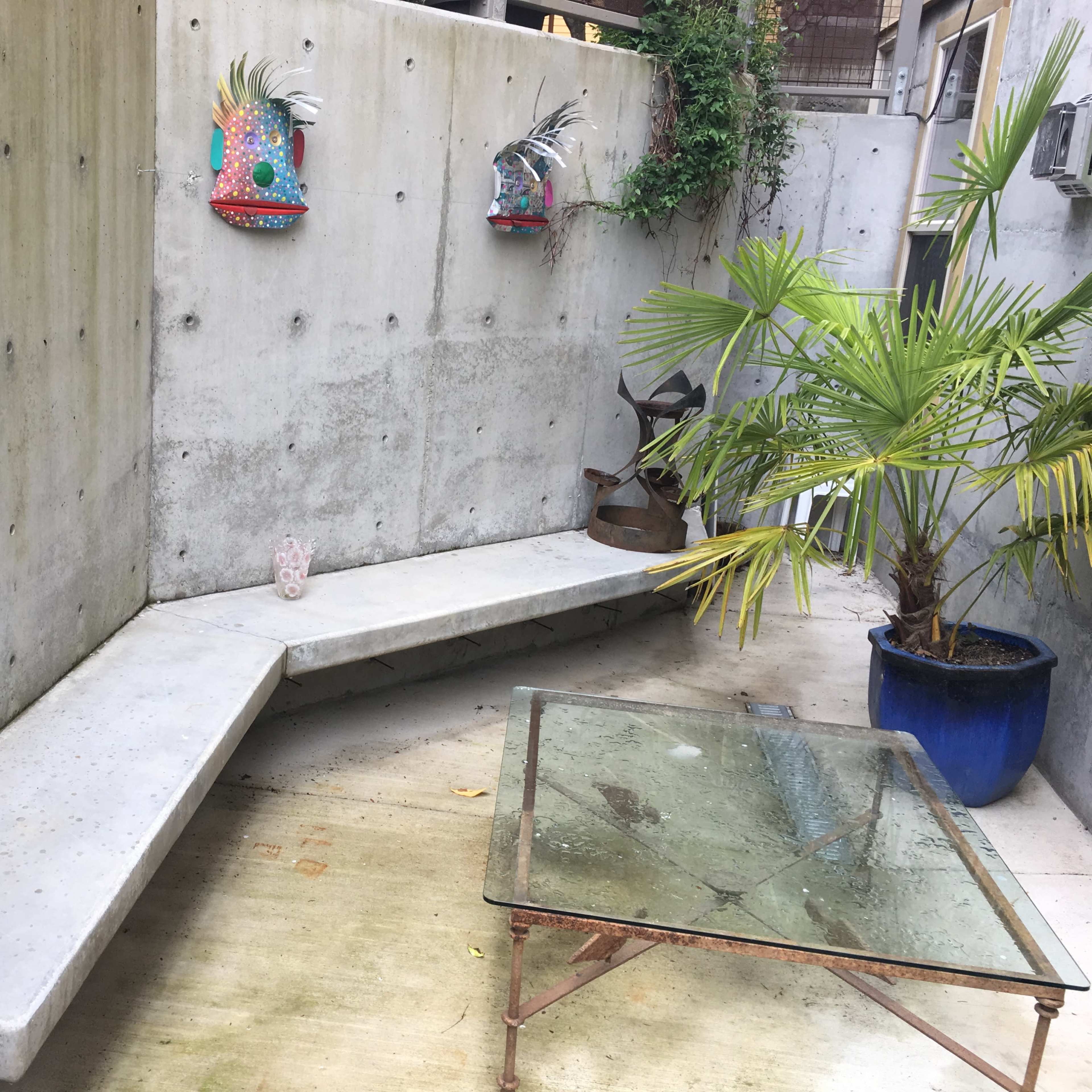 A small concrete courtyard features a curved bench and a glass-top table, with decorative wall art and potted plants.