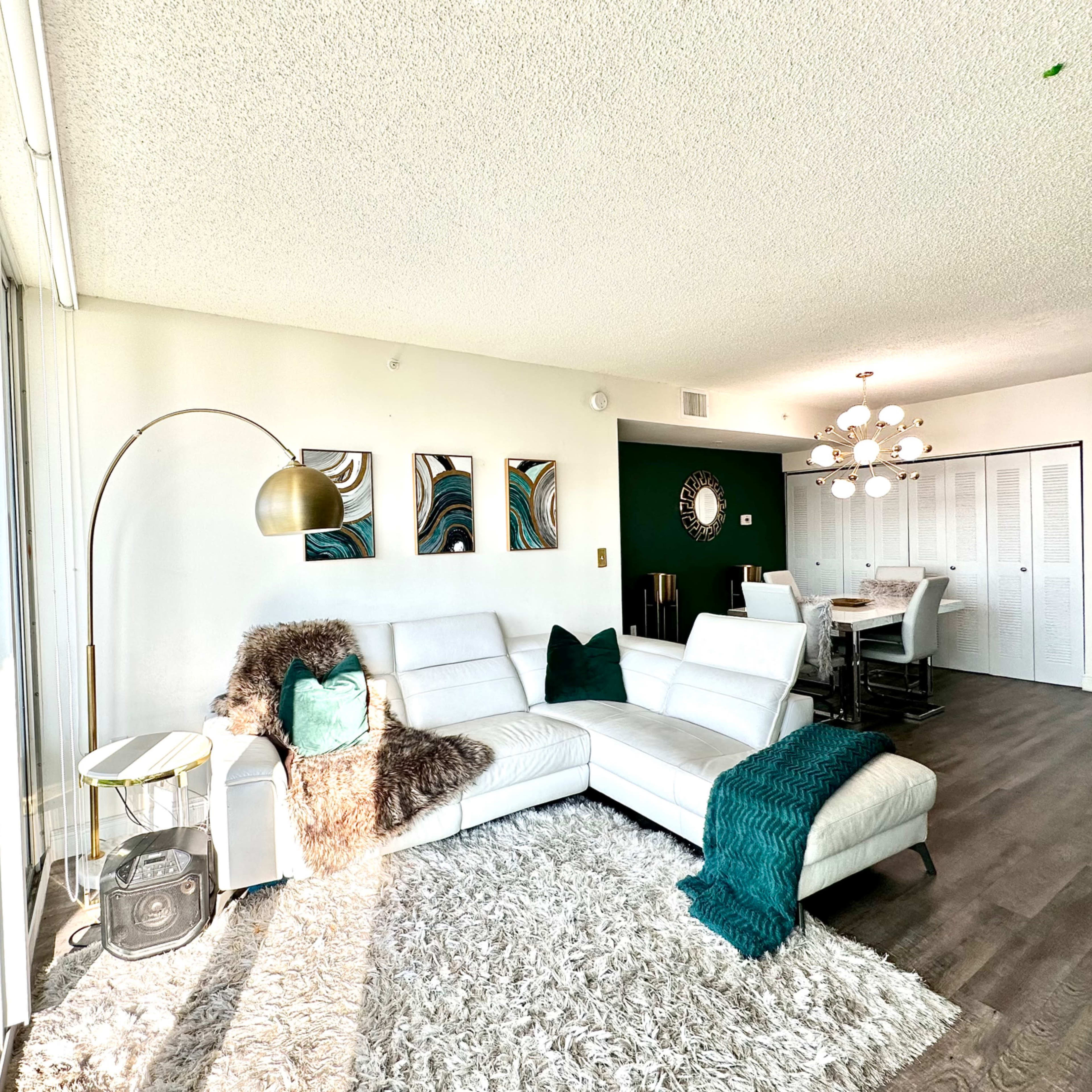 A modern living room features a white sectional sofa, a round floor lamp, and a textured rug, with artwork on the walls and a dining area in the background.